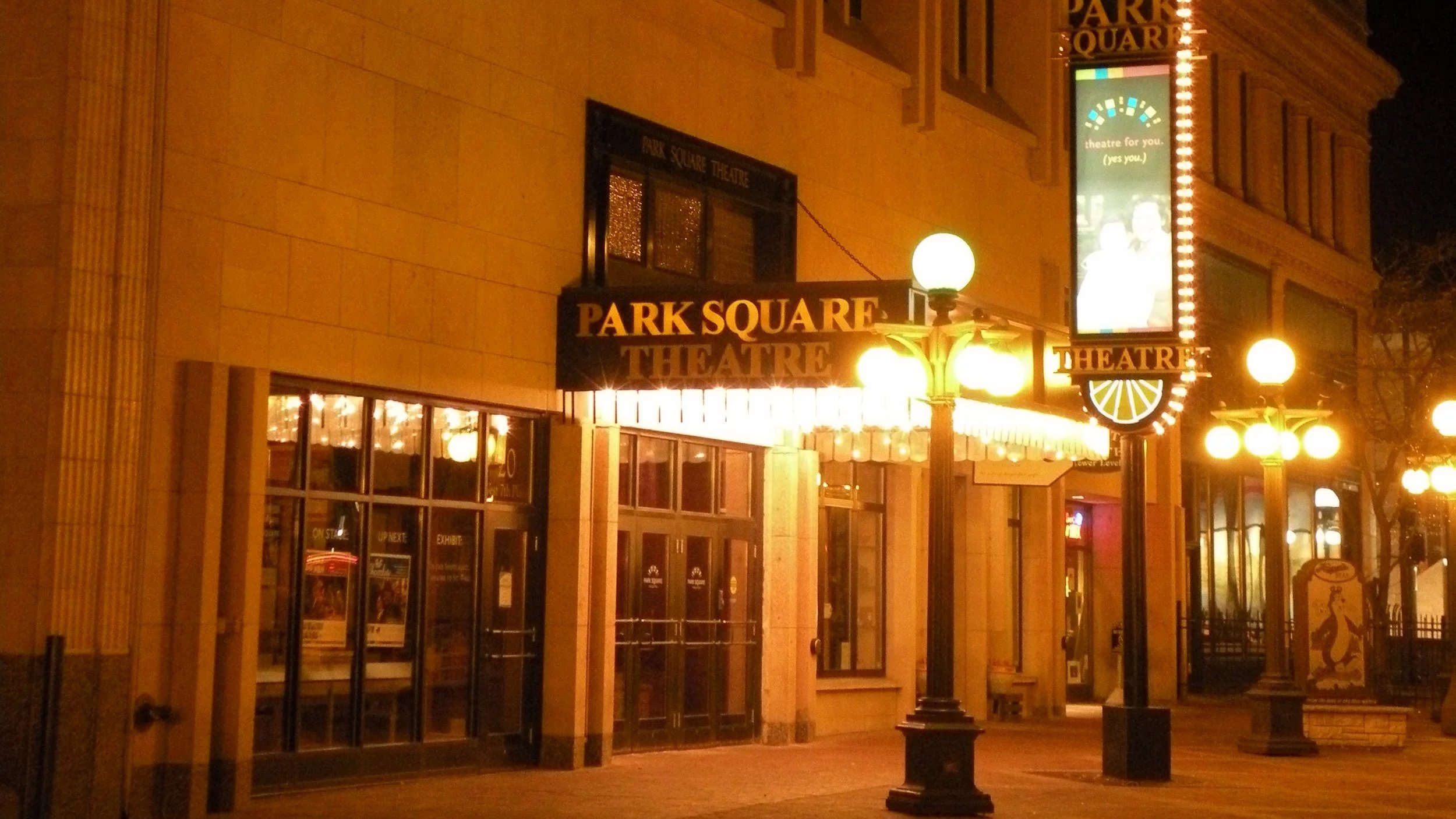 Night view of the illuminated Park Square Theatre with lit street lamps, a marquee sign, entrance doors, and a vertical digital display showing a scene and text.
