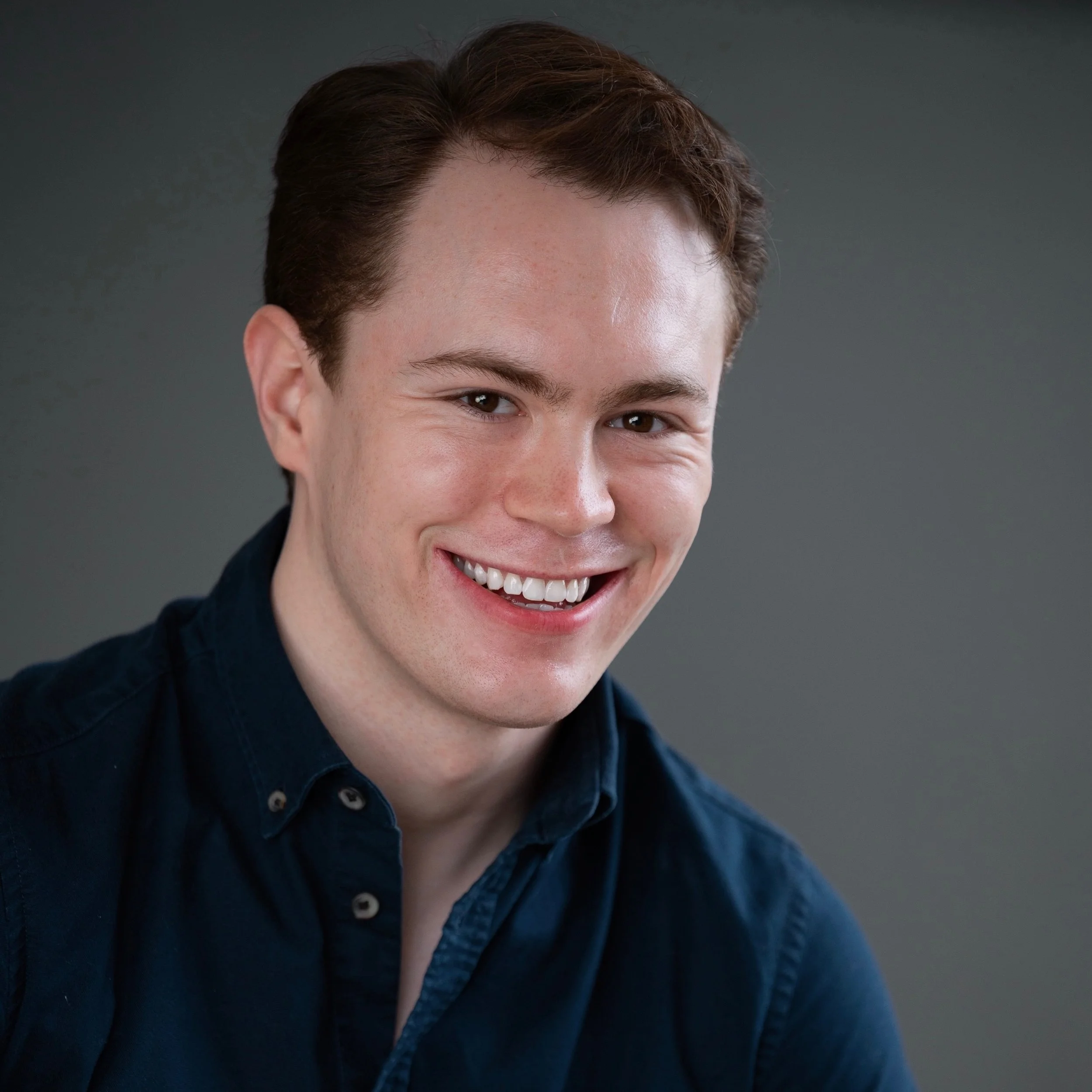 A young Caucasian man with short brown hair wearing a dark blue button-up shirt, smiling in front of a gray background.