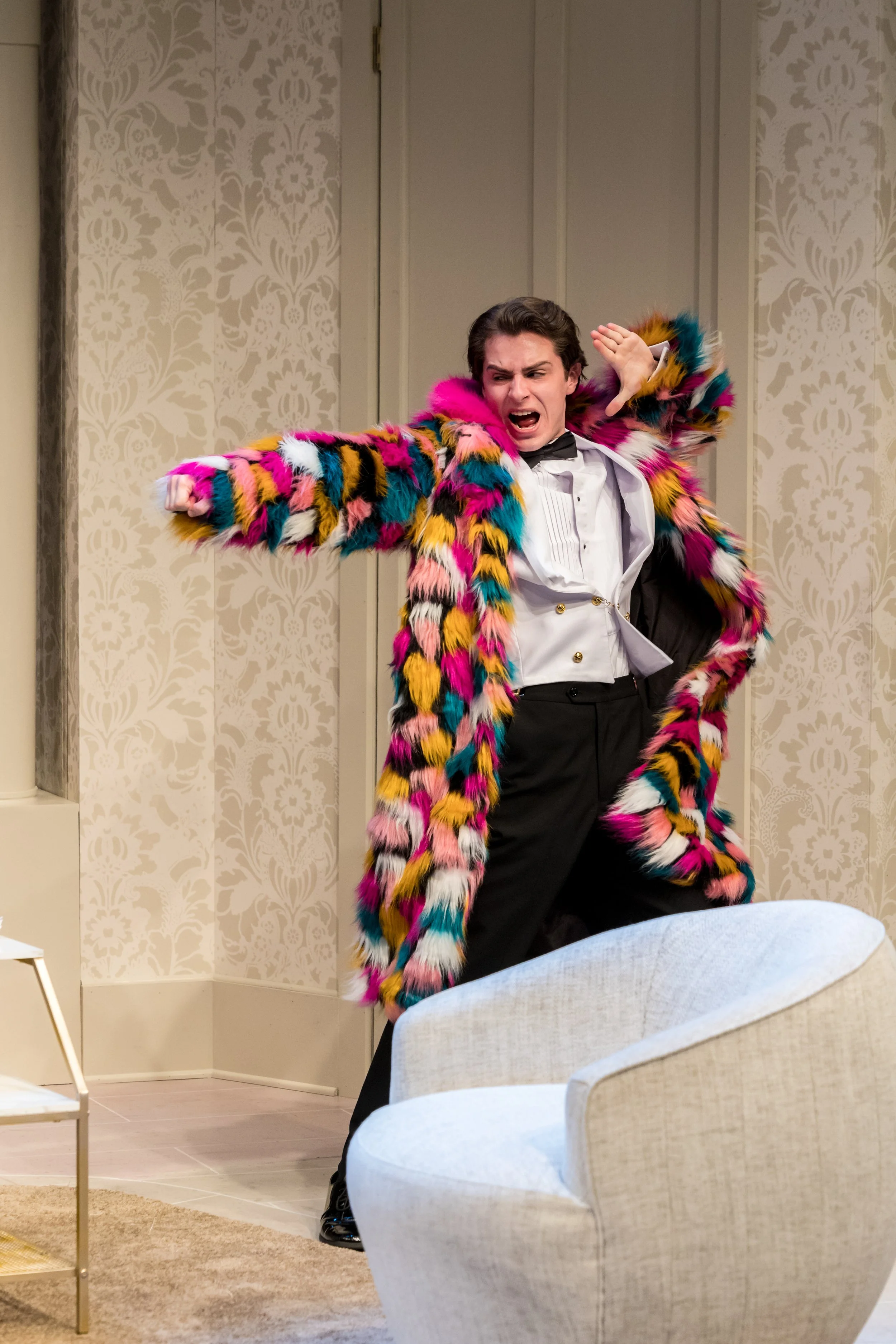 A man dressed in a colorful, furry, multicolored coat, white dress shirt, and black pants, appears to be escaping or pretending to run, with a dramatic expression on his face in a room with patterned wallpaper and contemporary furniture.