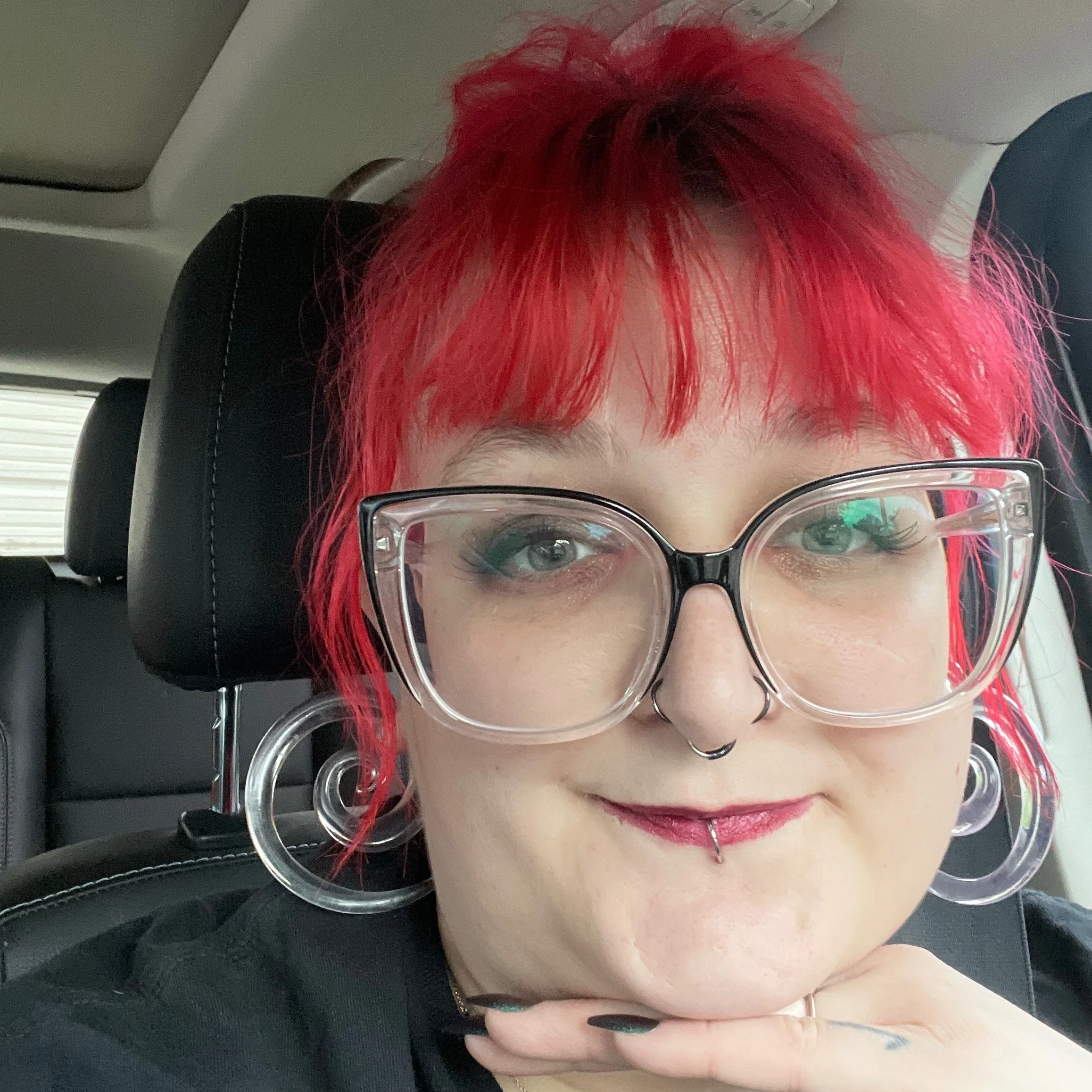 Person with bright red hair and large clear glasses, sitting in a vehicle, smiling with a septum piercing and pink lipstick.