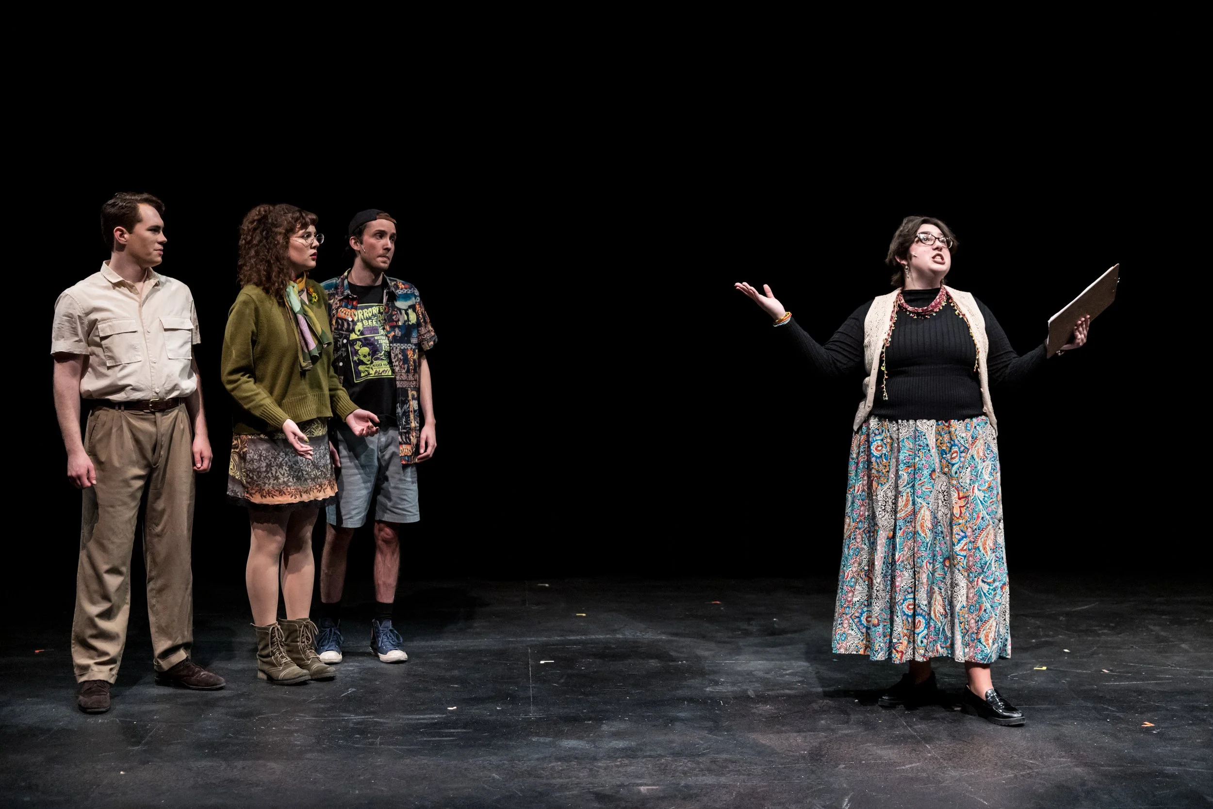 Four actors on stage, three listening attentively to a woman speaking, under dim lighting with a black background.