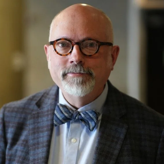 A middle-aged man with glasses, a beard and mustache, wearing a checkered blazer and a blue bow tie, appears to be in an indoor setting.