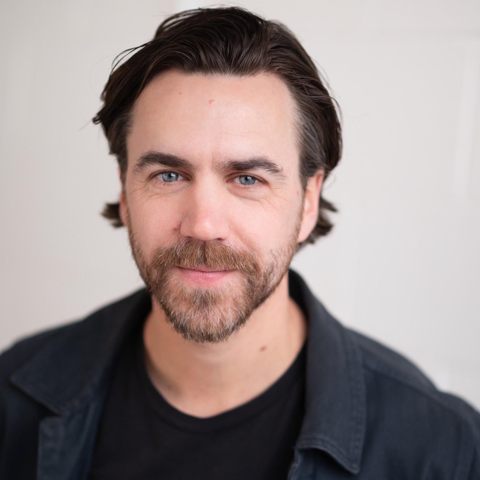 Close-up portrait of a man with shoulder-length dark hair, blue eyes, and a beard, wearing a black shirt and dark jacket contrast against a light background.