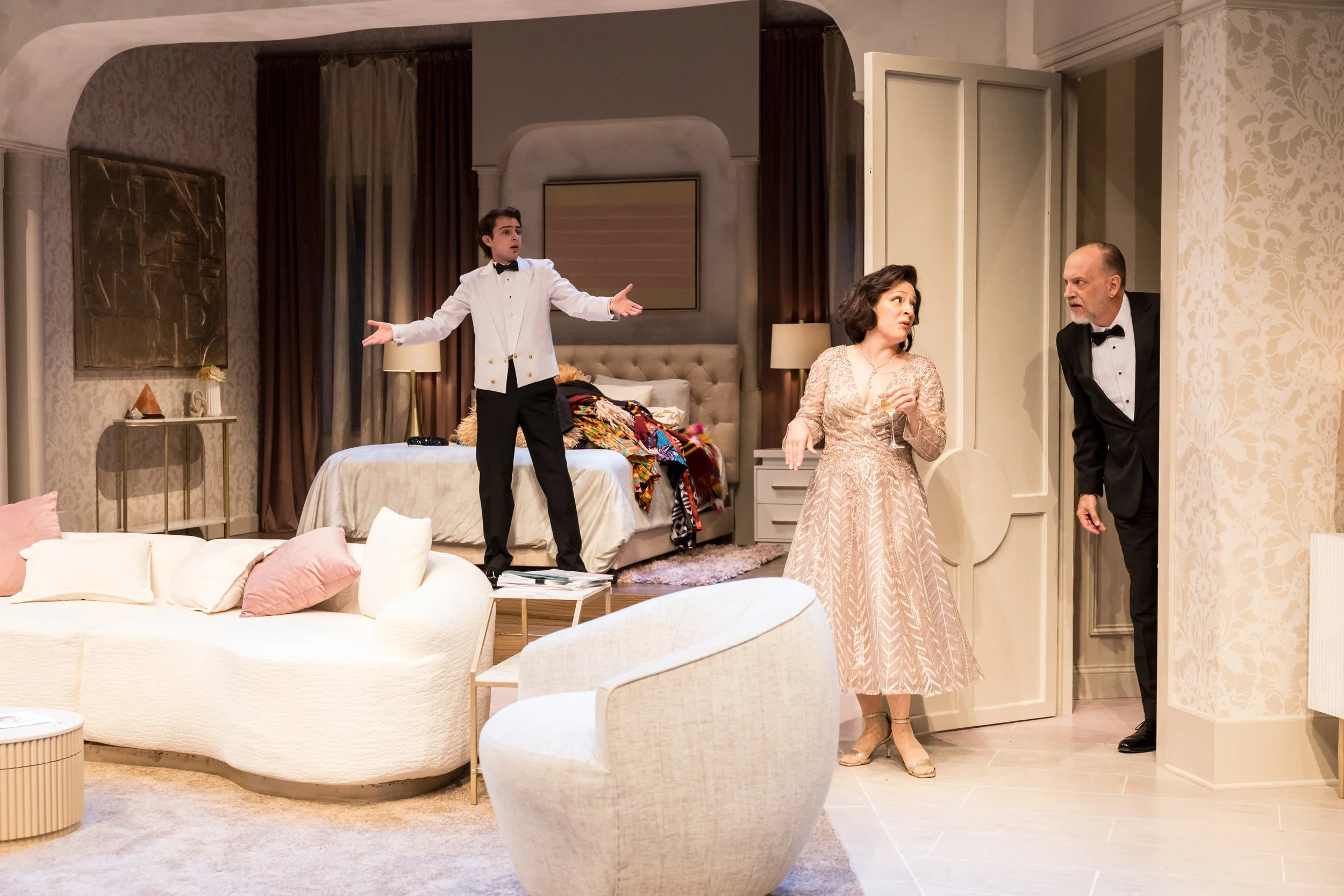 A scene from a play featuring three actors in a bedroom set: a young man in a white tuxedo with a black bow tie standing on a bed with open arms, a woman in a gold dress holding a glass of wine looking at an older man in a black tuxedo who is leaning