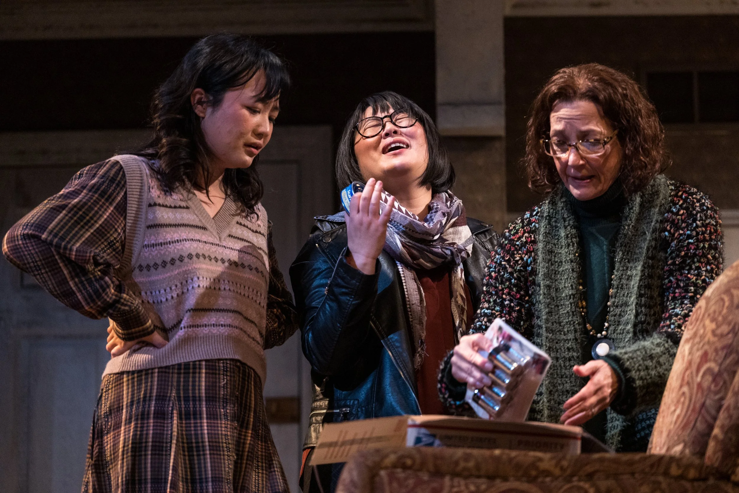 Three women standing around a table, looking at a small box, with one appearing emotional and another holding a small object, in a cozy indoor setting.