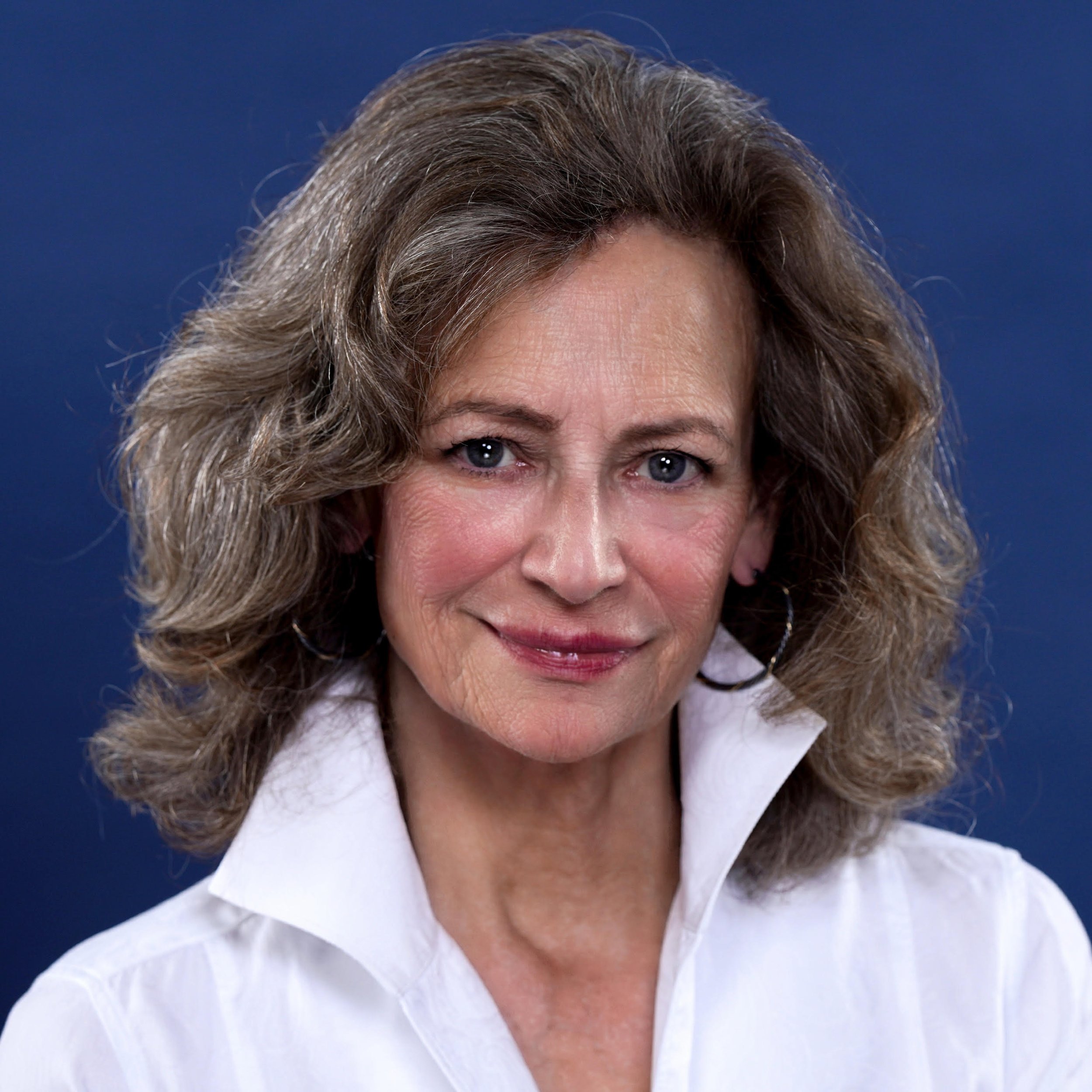 Portrait of a smiling middle-aged woman with curly brown hair, wearing a white collared shirt, set against a dark blue background.