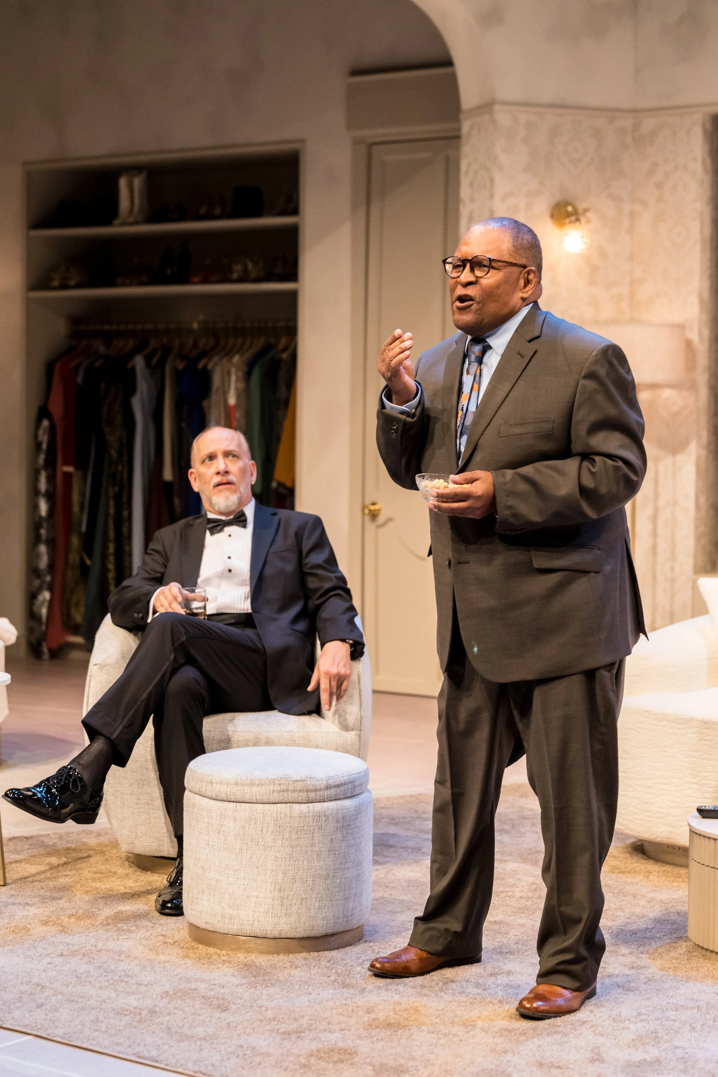 Two men in formal attire inside a room. One is seated, and the other is standing, holding a bowl and speaking. The seated man wears a tuxedo, and the standing man wears a suit and tie.