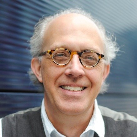 Close-up of a smiling man with glasses and curly gray hair, standing outdoors near a blue building.