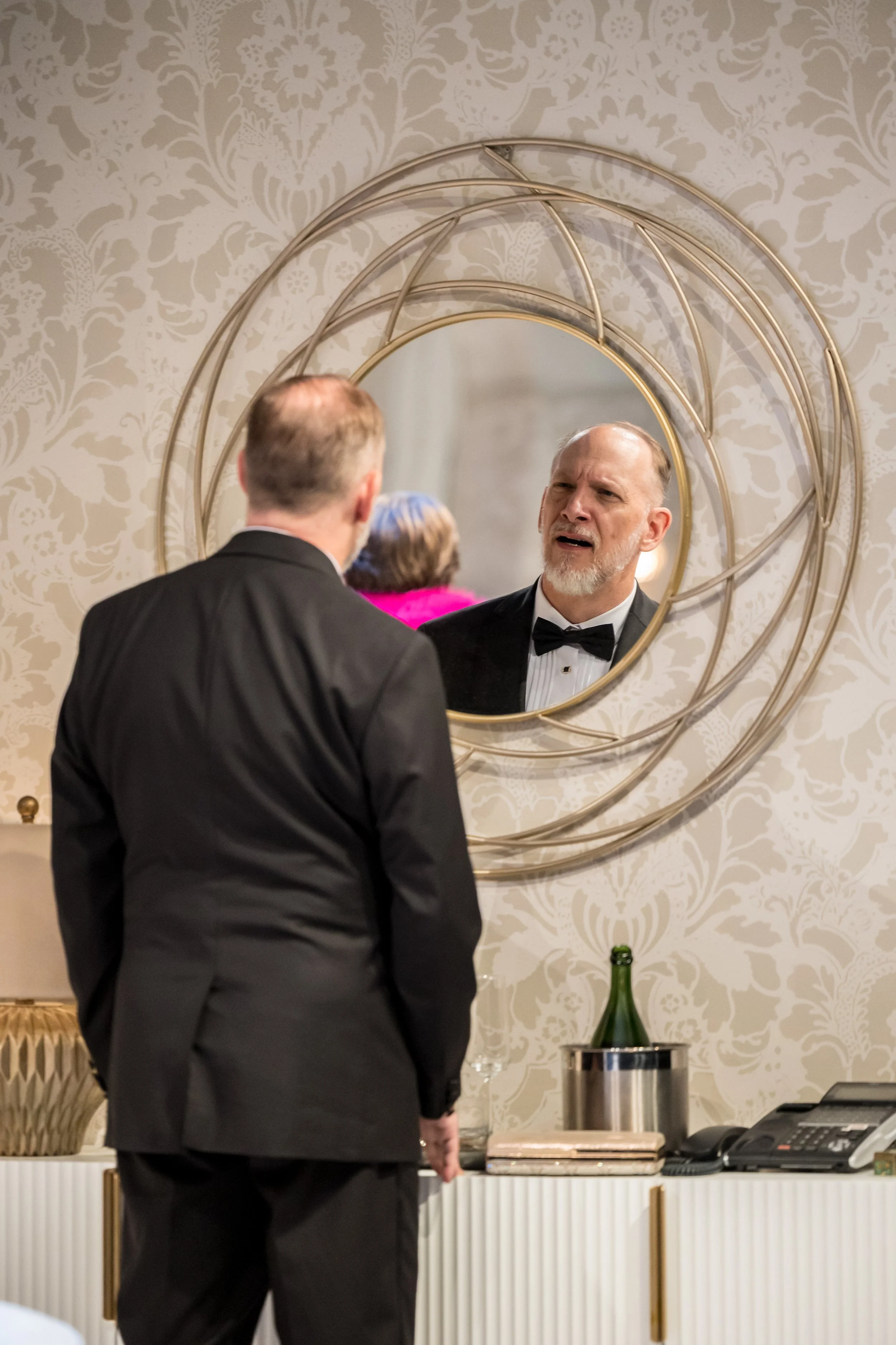 A man in a black tuxedo and bow tie looks at himself in a decorative round mirror on the wall, with a woman in a pink dress visible in the reflection.