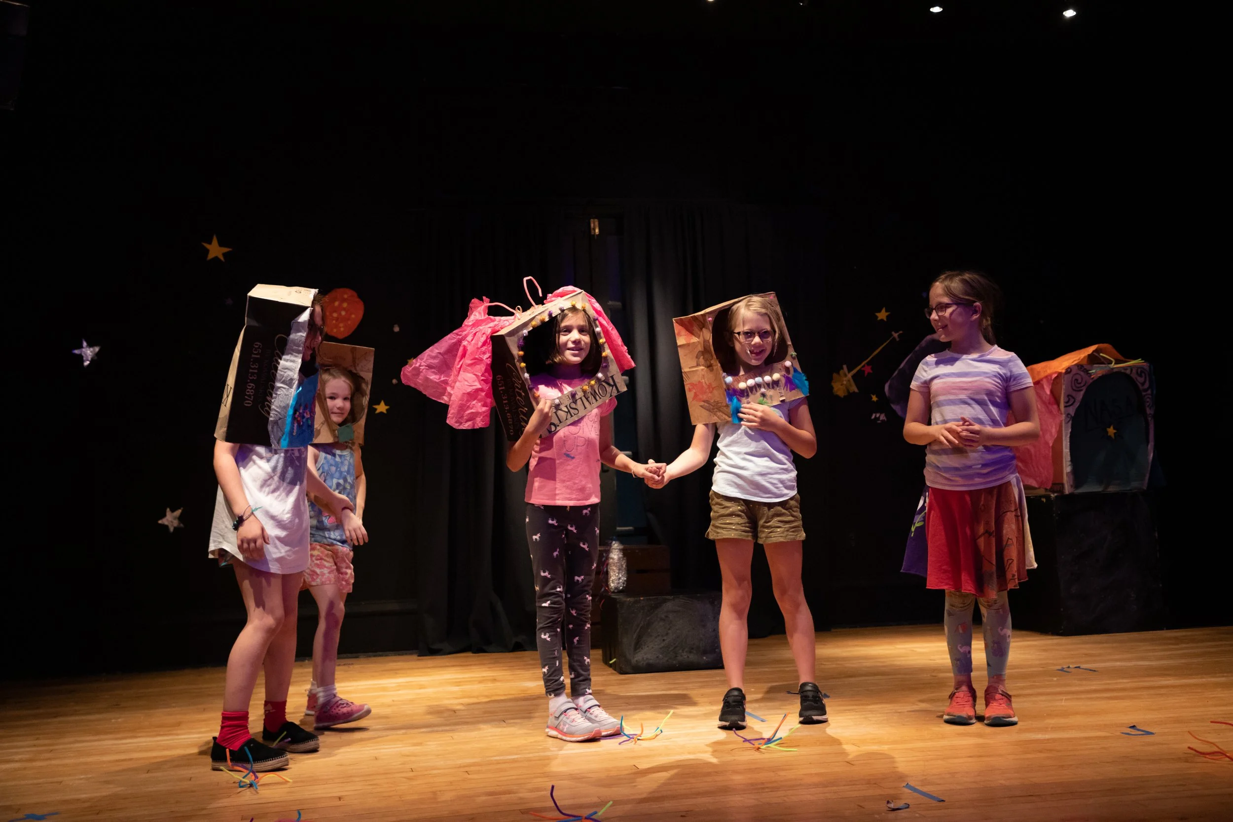 Five young girls on stage, wearing homemade costume boxes on their heads and holding hands, during a school play or performance with a black curtain background decorated with stars.