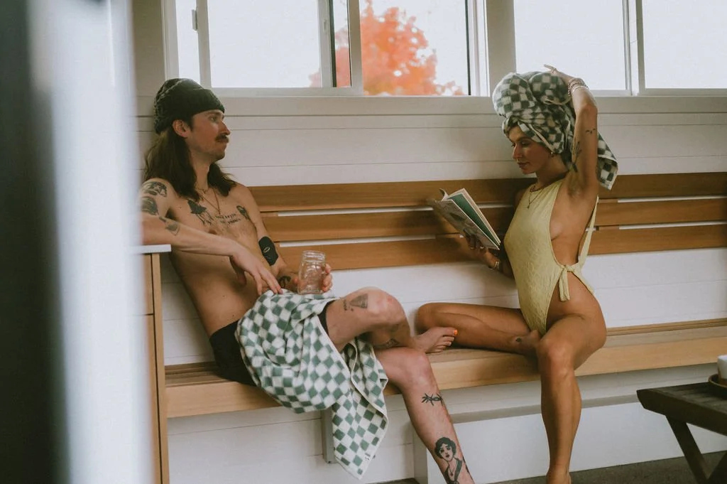 Tattooed man holding a drink and woman with towel-wrapped hair reading a magazine in a bright Nordica Haus lounge—modern Hood River sauna culture.