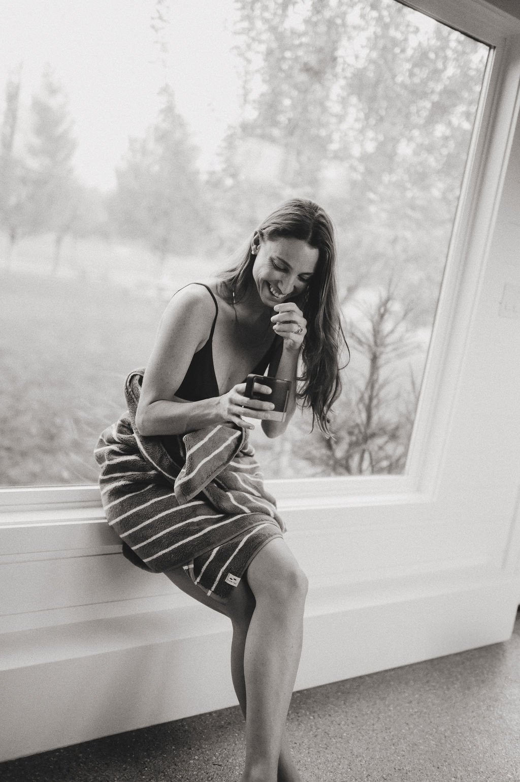 Woman wrapped in a striped towel sitting on a window sill at Nordica Haus, smiling at her phone with riverfront nature views behind her.