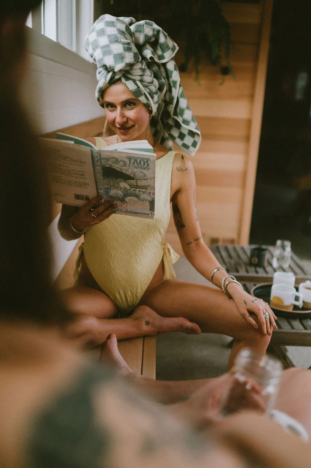 Woman with a towel on her head reading a book in the Nordica Haus wooden lounge, smiling into the camera—slow living and waterfront sauna vibes.