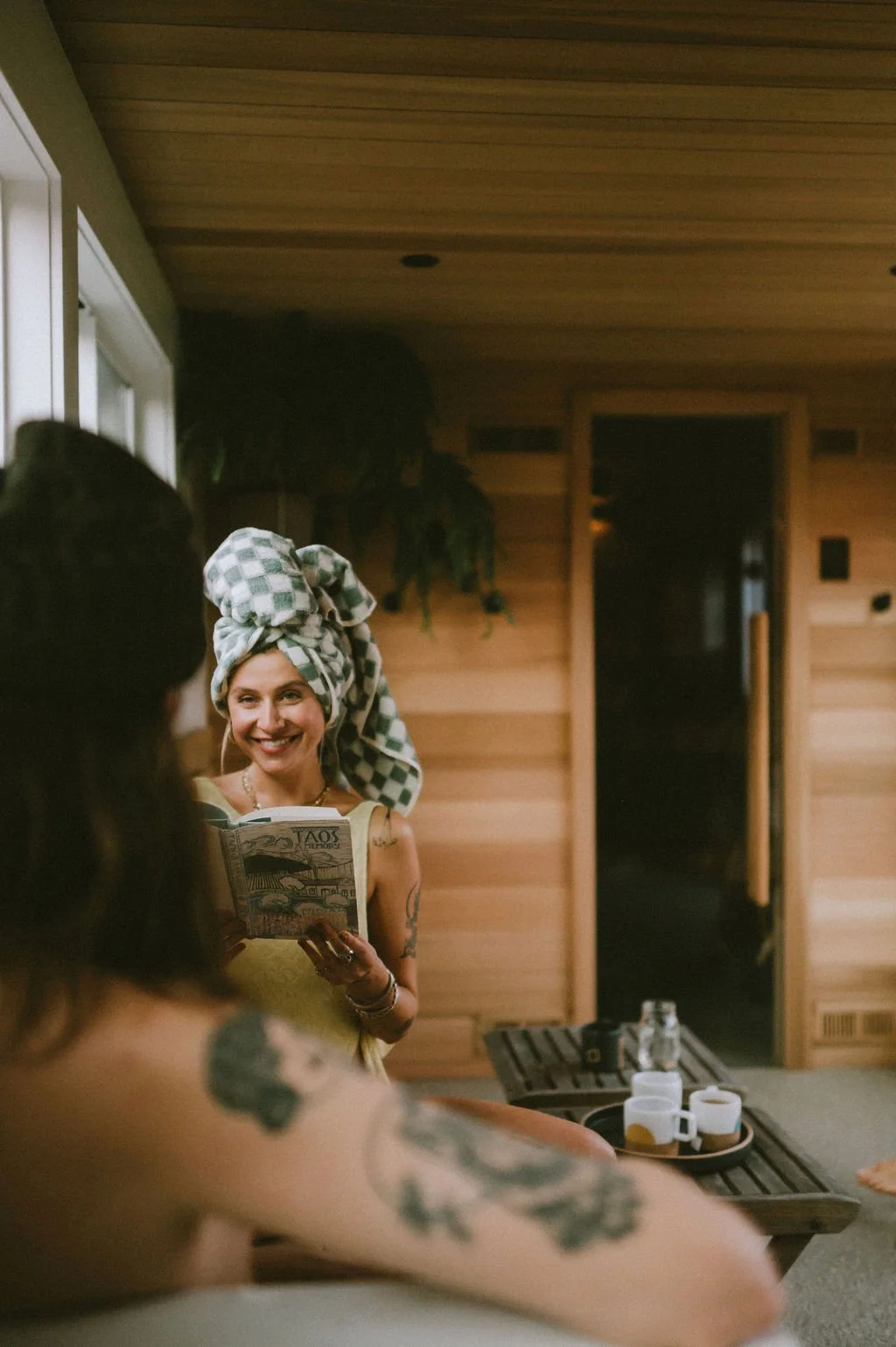 Woman with a towel-wrapped head smiling with a booklet inside the Nordica Haus sauna lounge, surrounded by warm wood, friends, and cozy Hood River wellness vibes.