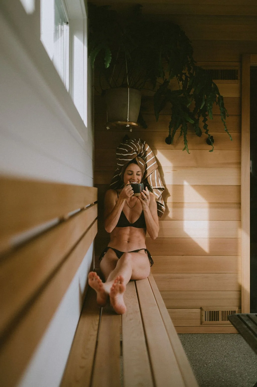 Woman in a black bikini with a towel-wrapped head sitting in the Nordica Haus sauna—warm wood, hanging plants, and serene heat therapy.