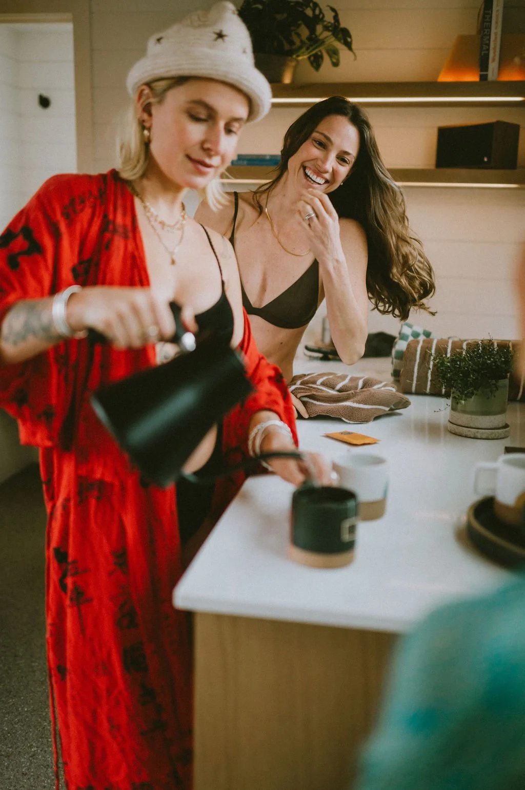 Two women in lingerie sharing drinks and laughter in a cozy Nordica Haus kitchen space—playful energy meets modern Hood River wellness culture.