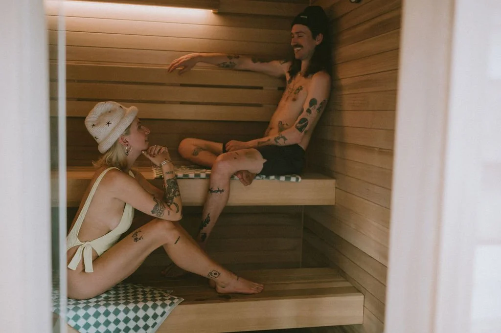 Tattooed man and woman sitting on wooden benches inside the Nordica Haus sauna—heat, connection, and modern Scandinavian wellness style.