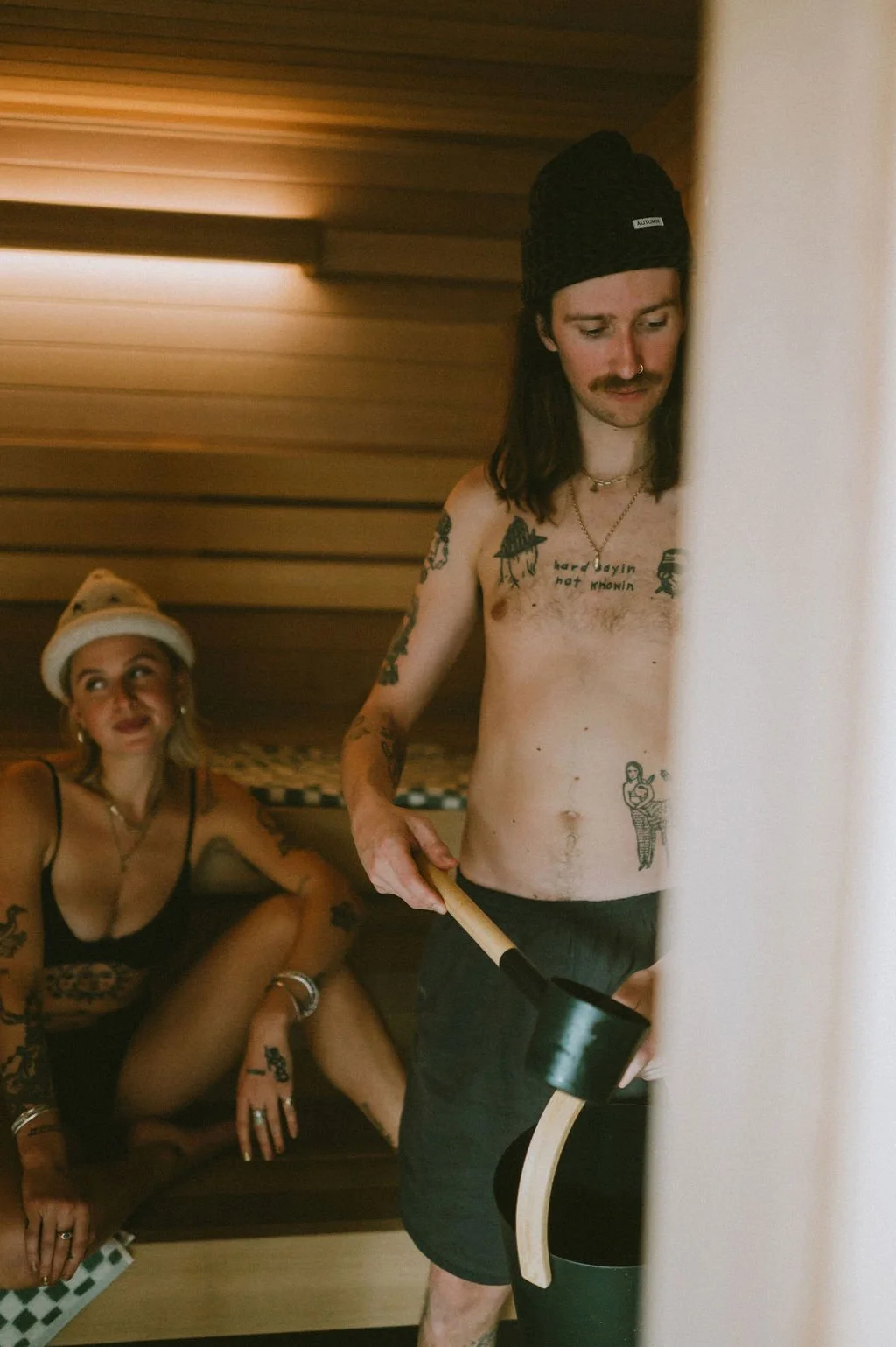 Tattooed man with a beanie holding a sauna ladle inside Nordica Haus, with a relaxed woman in a white hat behind him—heat, attitude, and Scandinavian style.