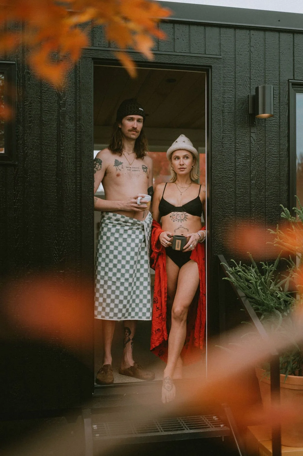 Tattooed man in a towel and woman in a hat and robe standing outside the Nordica Haus sauna—post-heat glow and effortless Scandinavian-cool vibes.