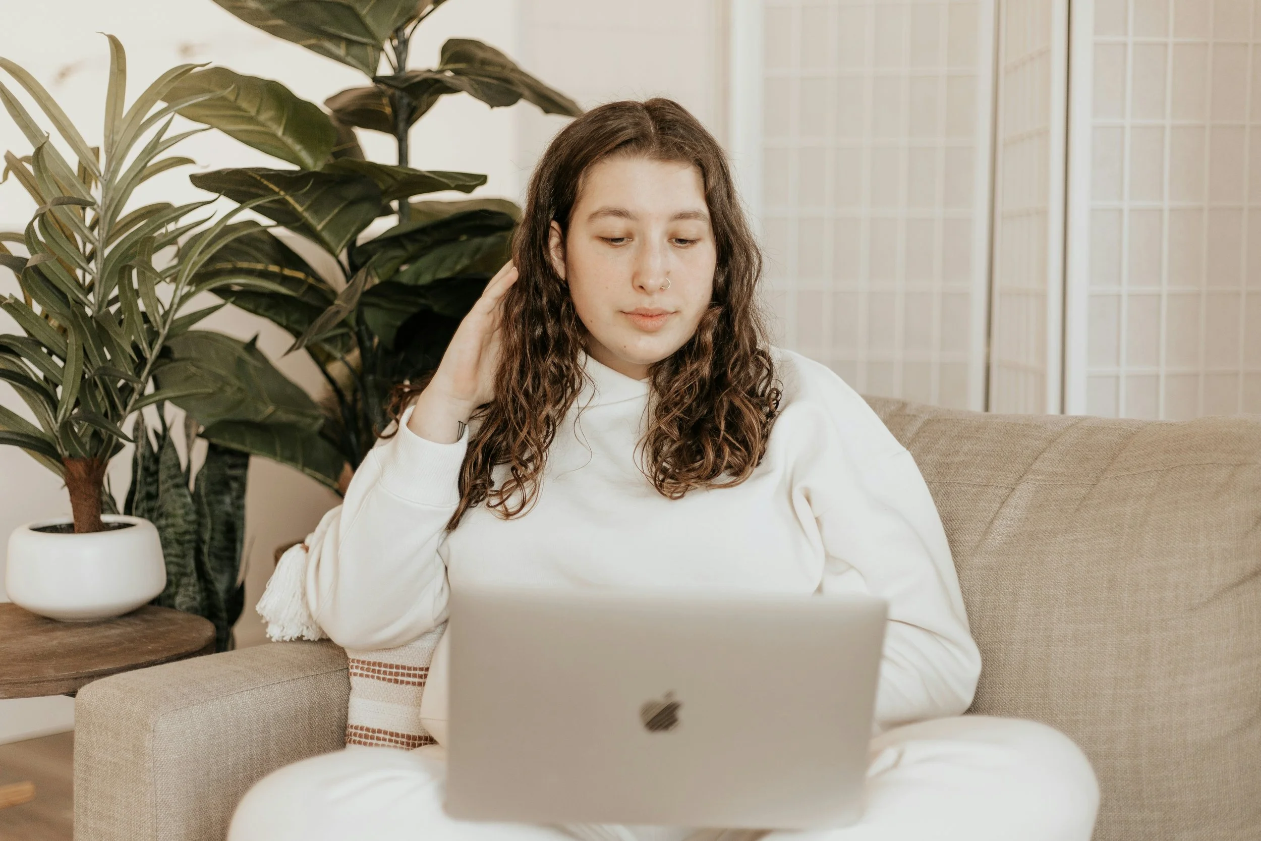 A young woman with wavy brown hair and a nose piercing sitting on a beige sofa, looking at a silver laptop on her lap, in a room with large green houseplants and a folding privacy screen in the background.