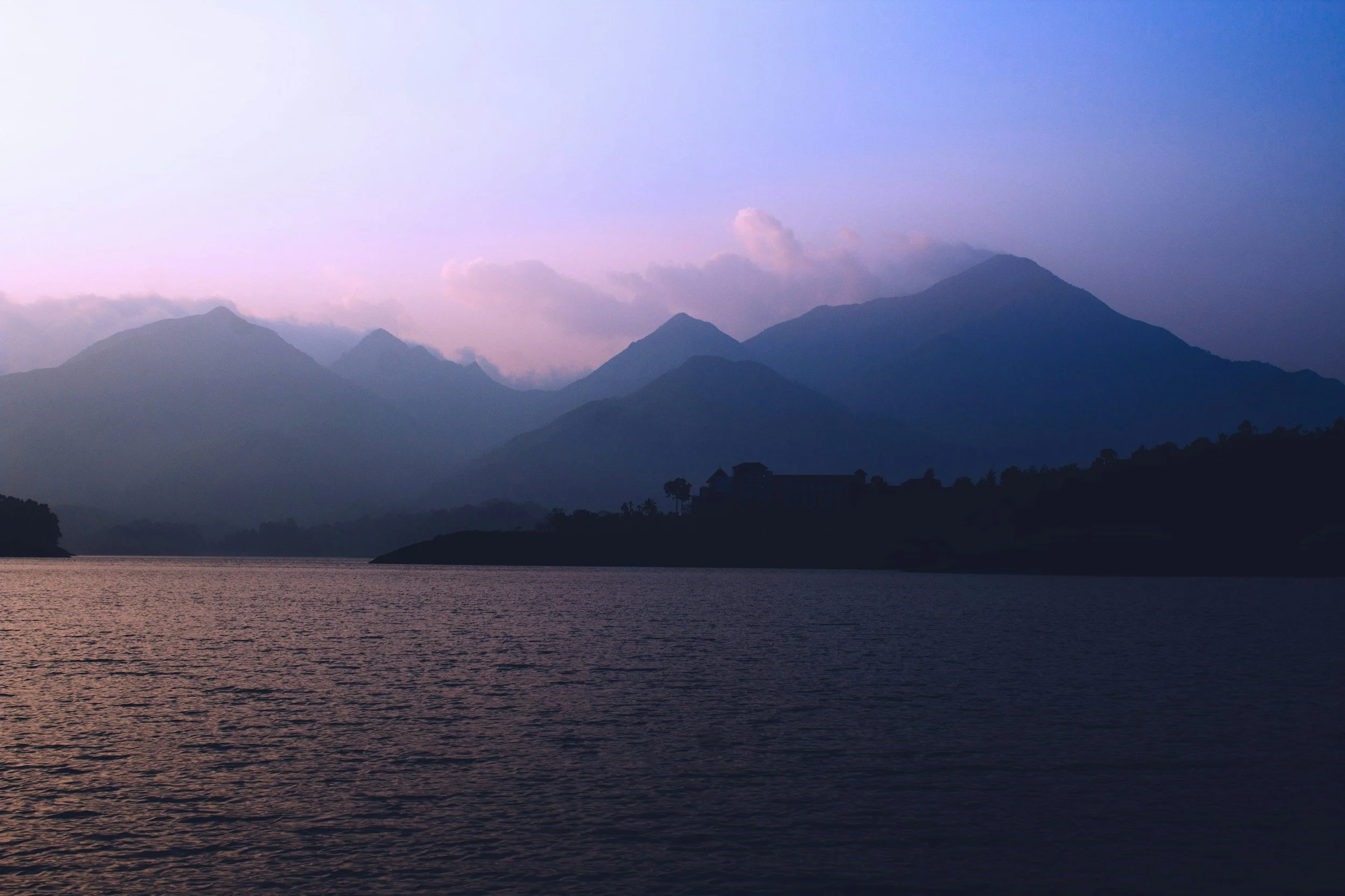 A tranquil scene of mountains at dusk with a lake in the foreground and clouds above.