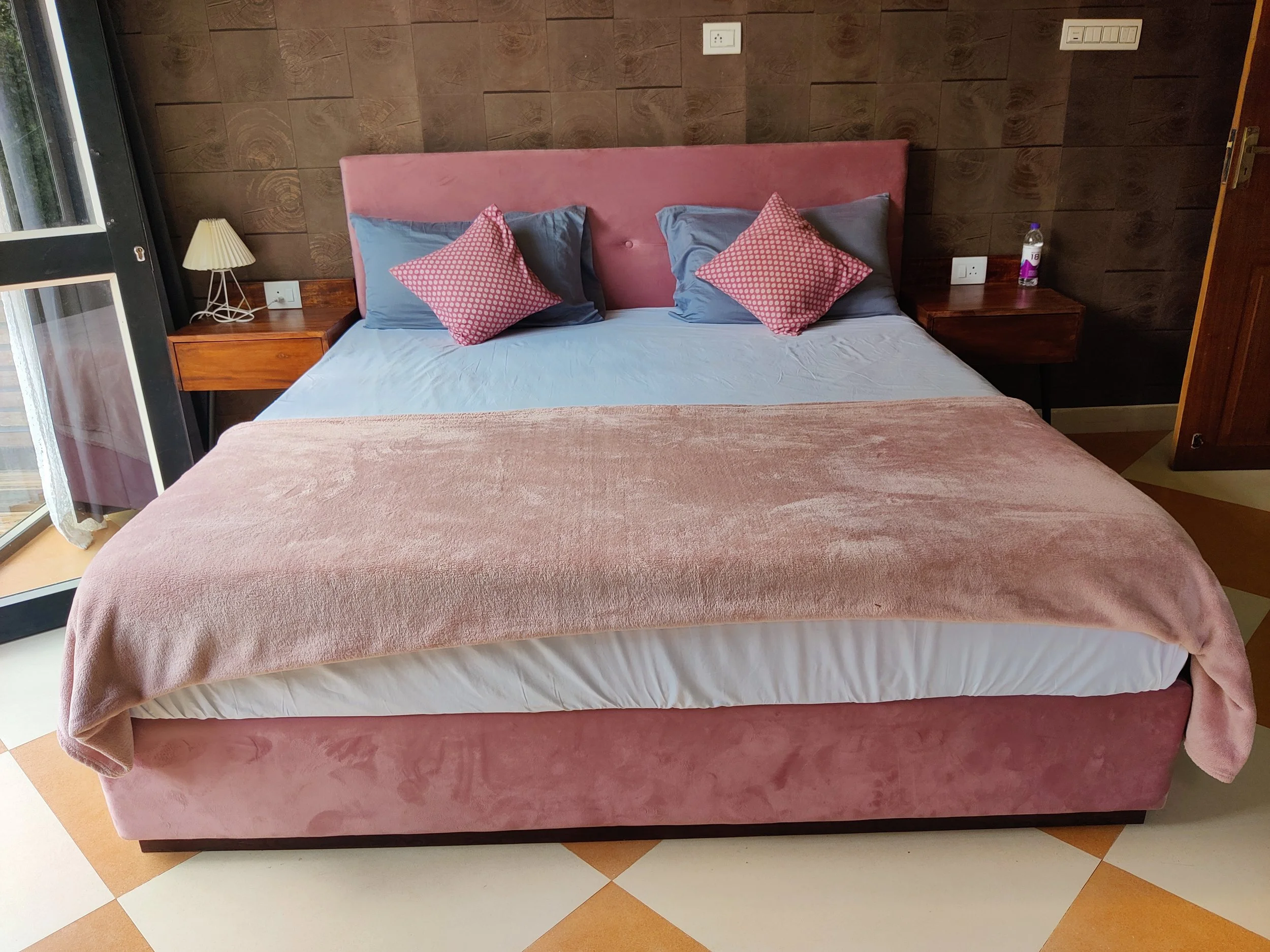 A neatly made bed with pink velvet headboard, gray and pink pillows, a pink blanket, and two wooden side tables with a lamp and water bottle in a bedroom with dark brown wood paneling.