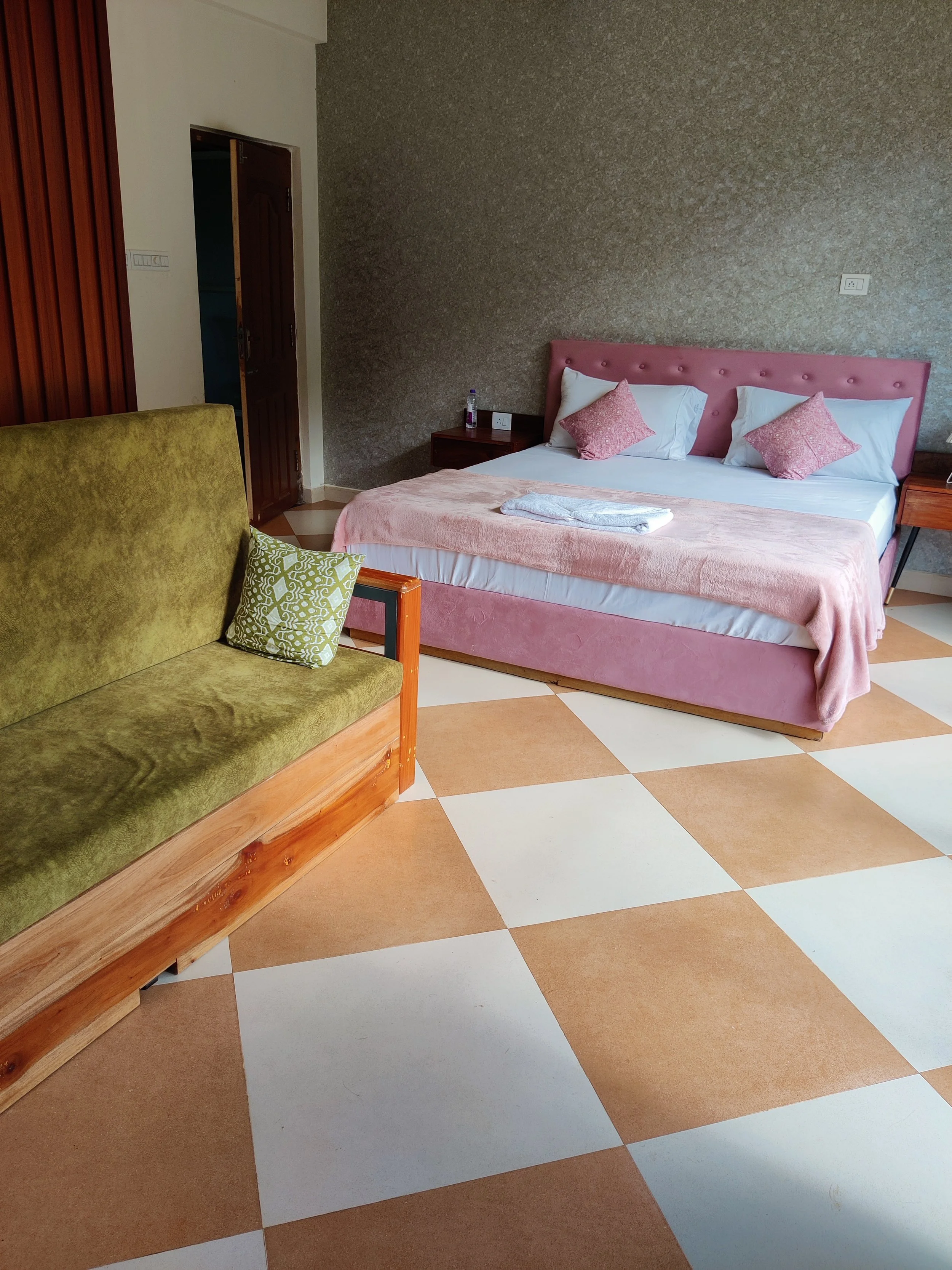 A hotel room with a pink upholstered bed, a wooden side table, and a green velvet sofa with a decorative pillow. The floor has large beige and white checkered tiles.