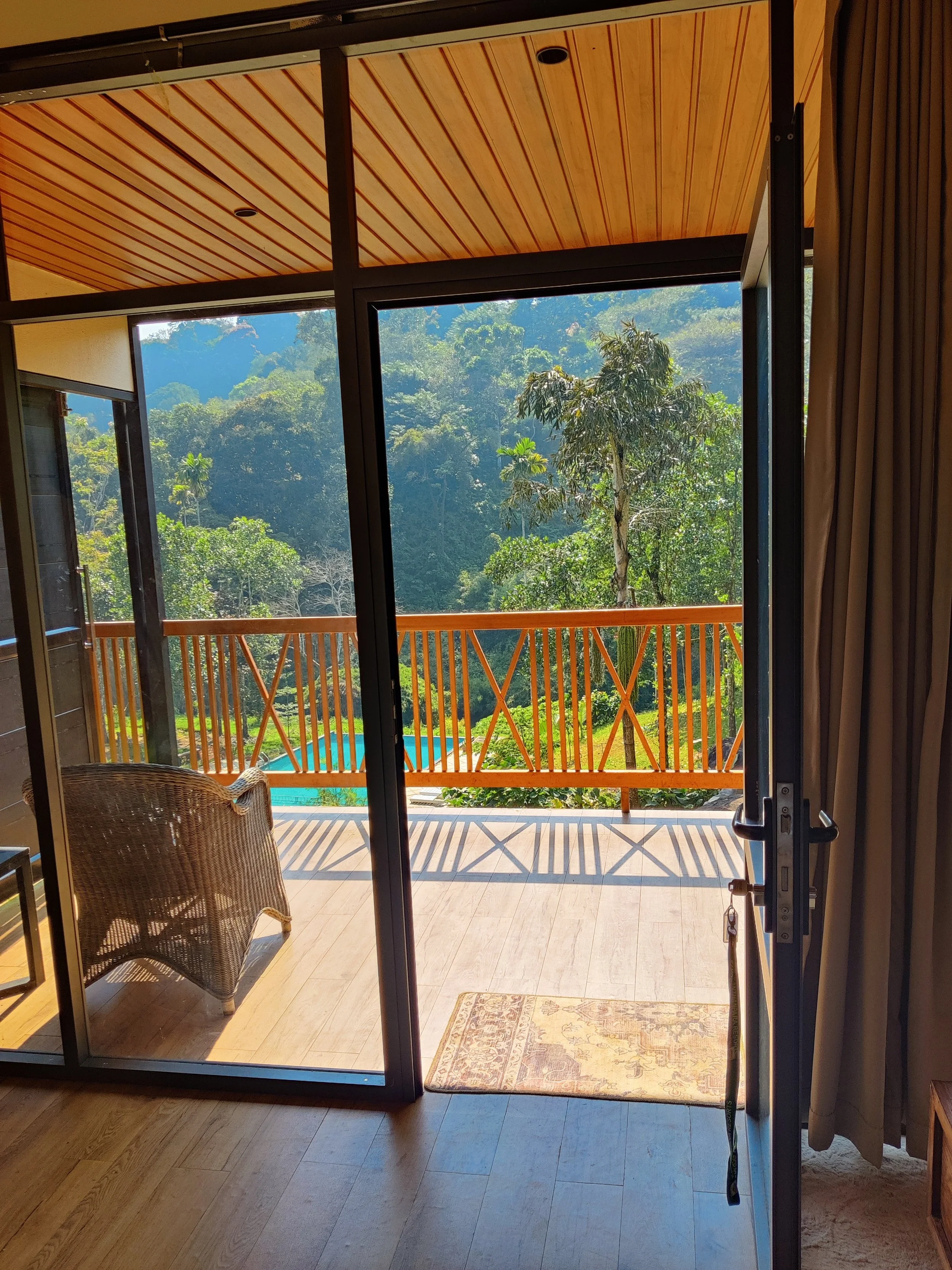 View from inside a room looking out onto a wooden balcony with outdoor furniture and lush green trees and mountains in the background.