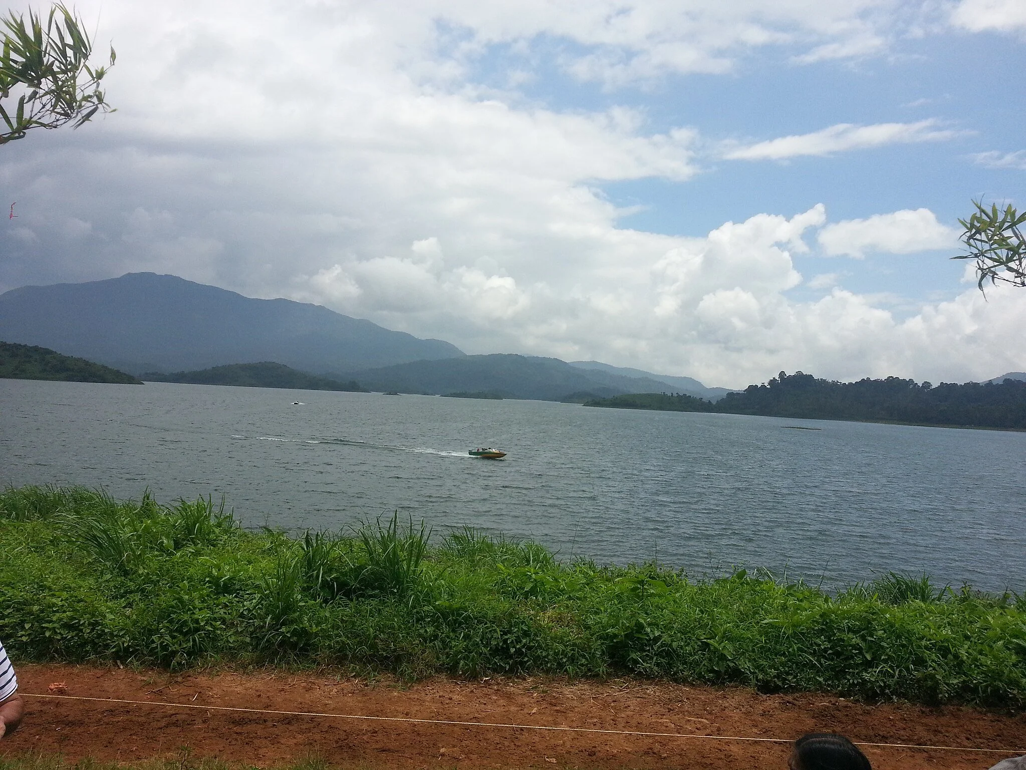 Scenic view of a lake with a boat, surrounded by mountains and partly cloudy sky, with green grass and dirt in the foreground.
