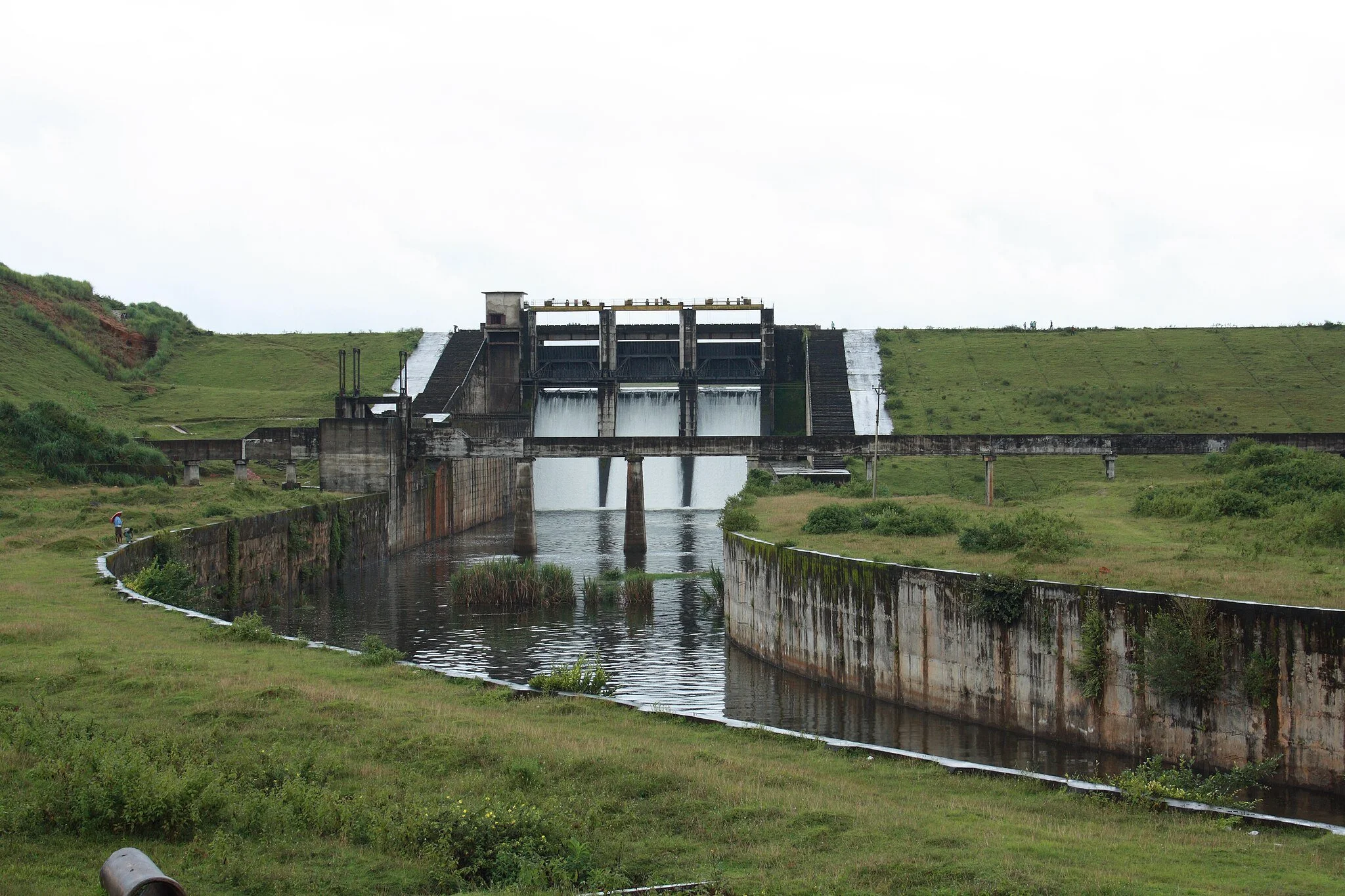 A dam with water flowing through spillways, surrounded by green grassy hills under a cloudy sky.