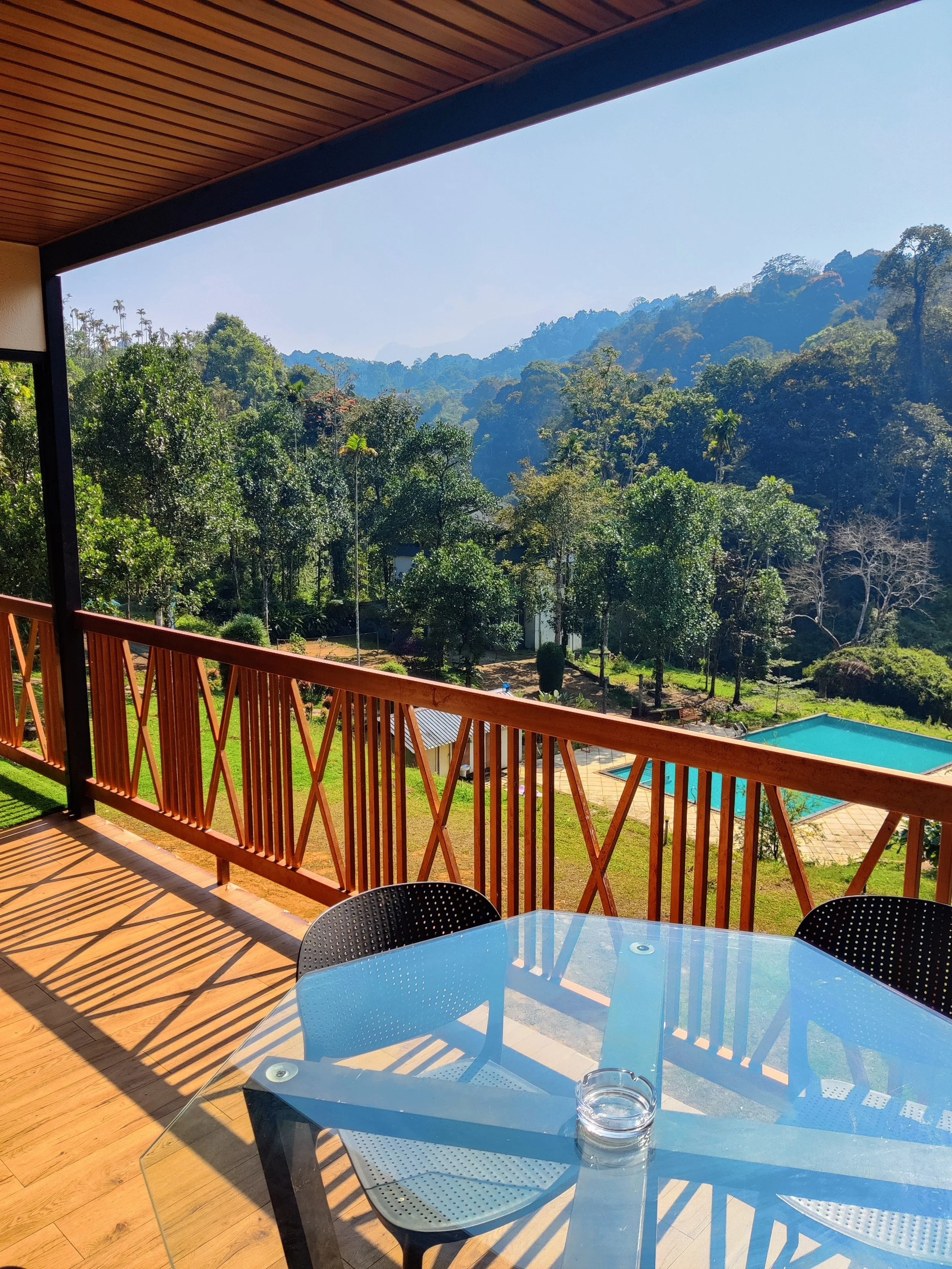 Balcony overlooking a lush green forest with mountains in the background, near a swimming pool, with a glass-topped table and black chairs.
