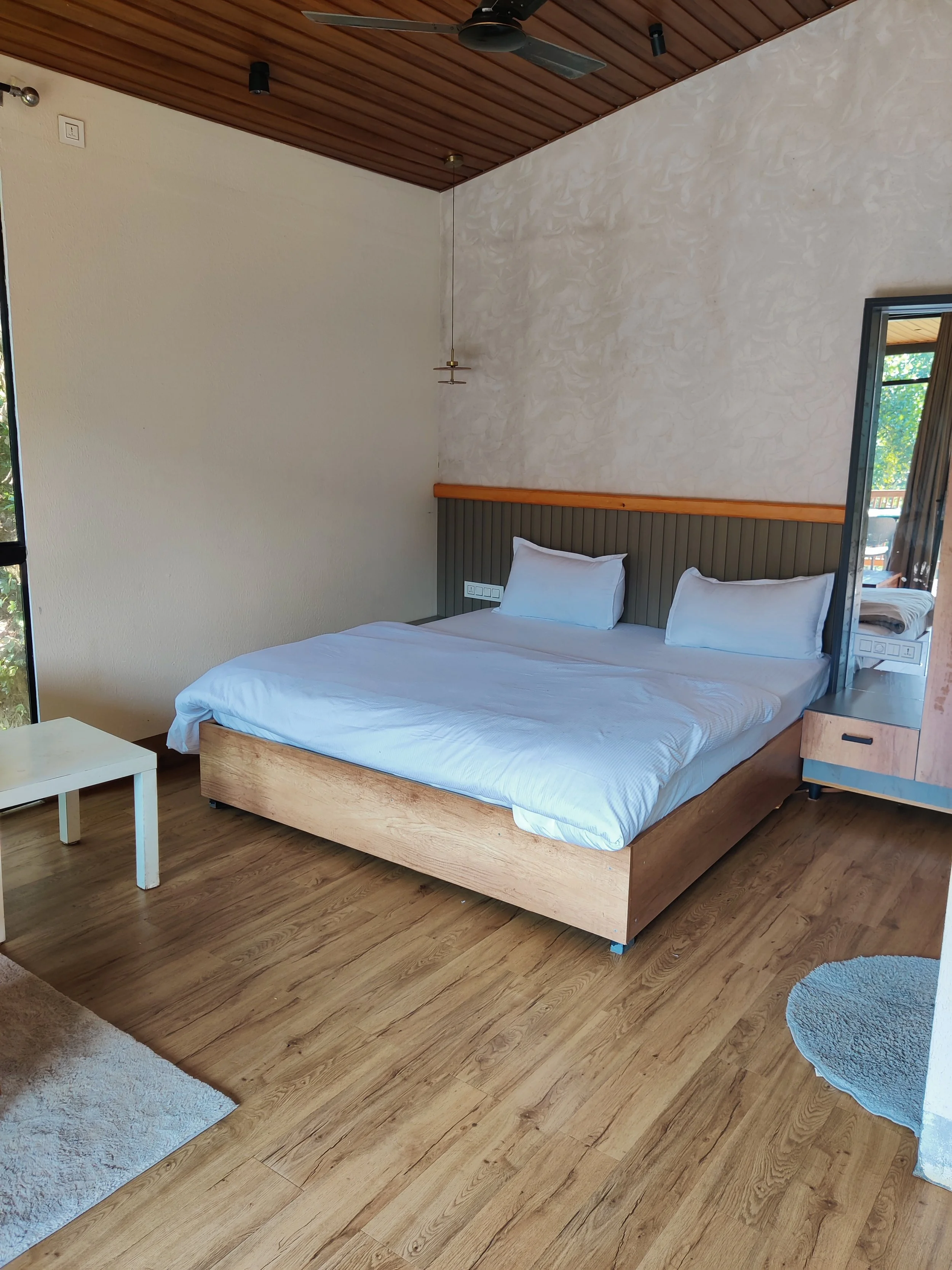 A bedroom with a wooden bed frame, white bedding, two pillows, a small white side table, a mirror on the wall, a wooden ceiling fan, and a sliding door leading to a balcony.