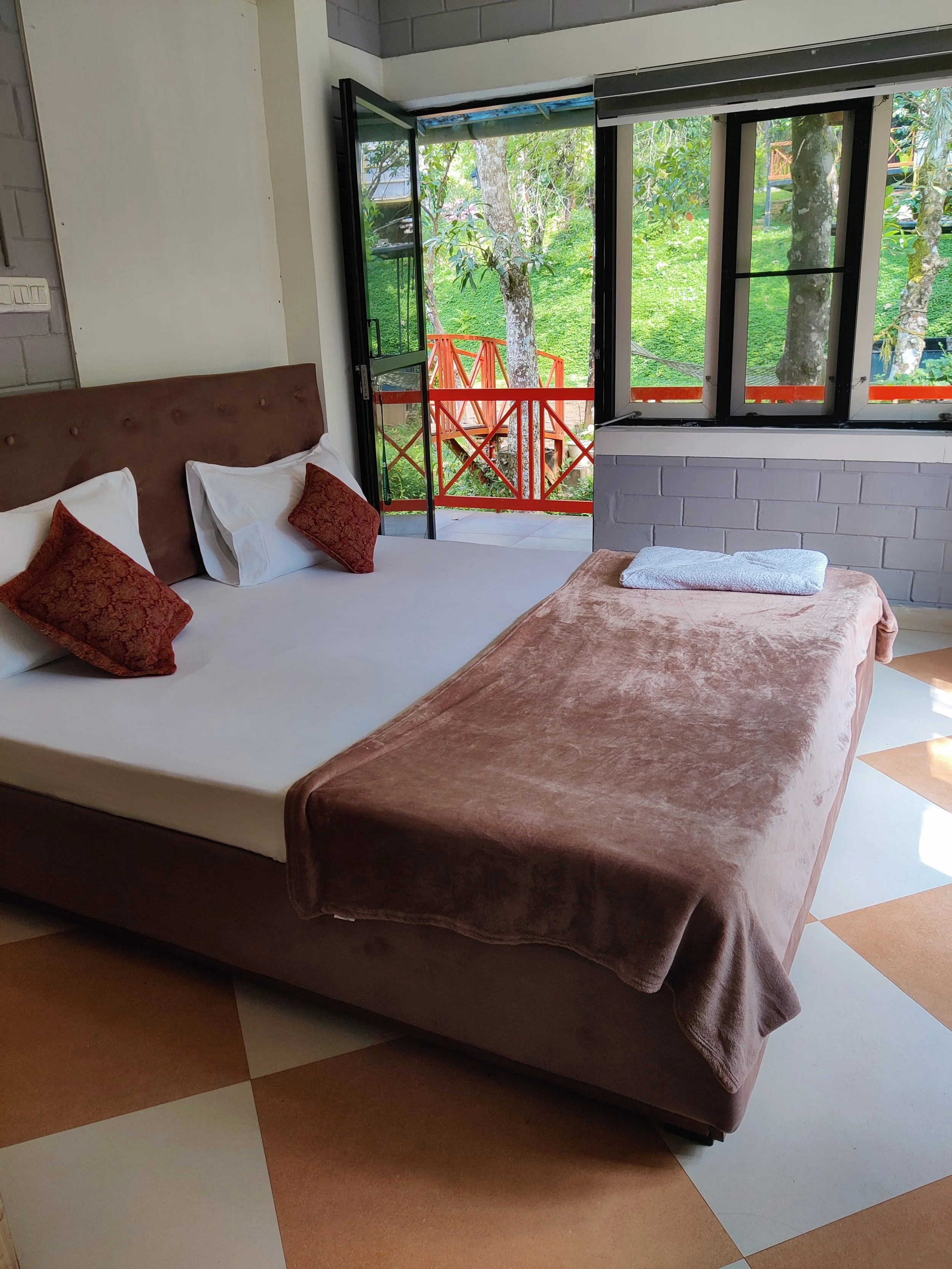 A bedroom with a large bed, made with white sheets and two white pillows, two smaller decorative orange pillows, a brown headboard, a folded towel on top of a brown blanket, and an open door leading to a balcony with a view of green trees and a red r
