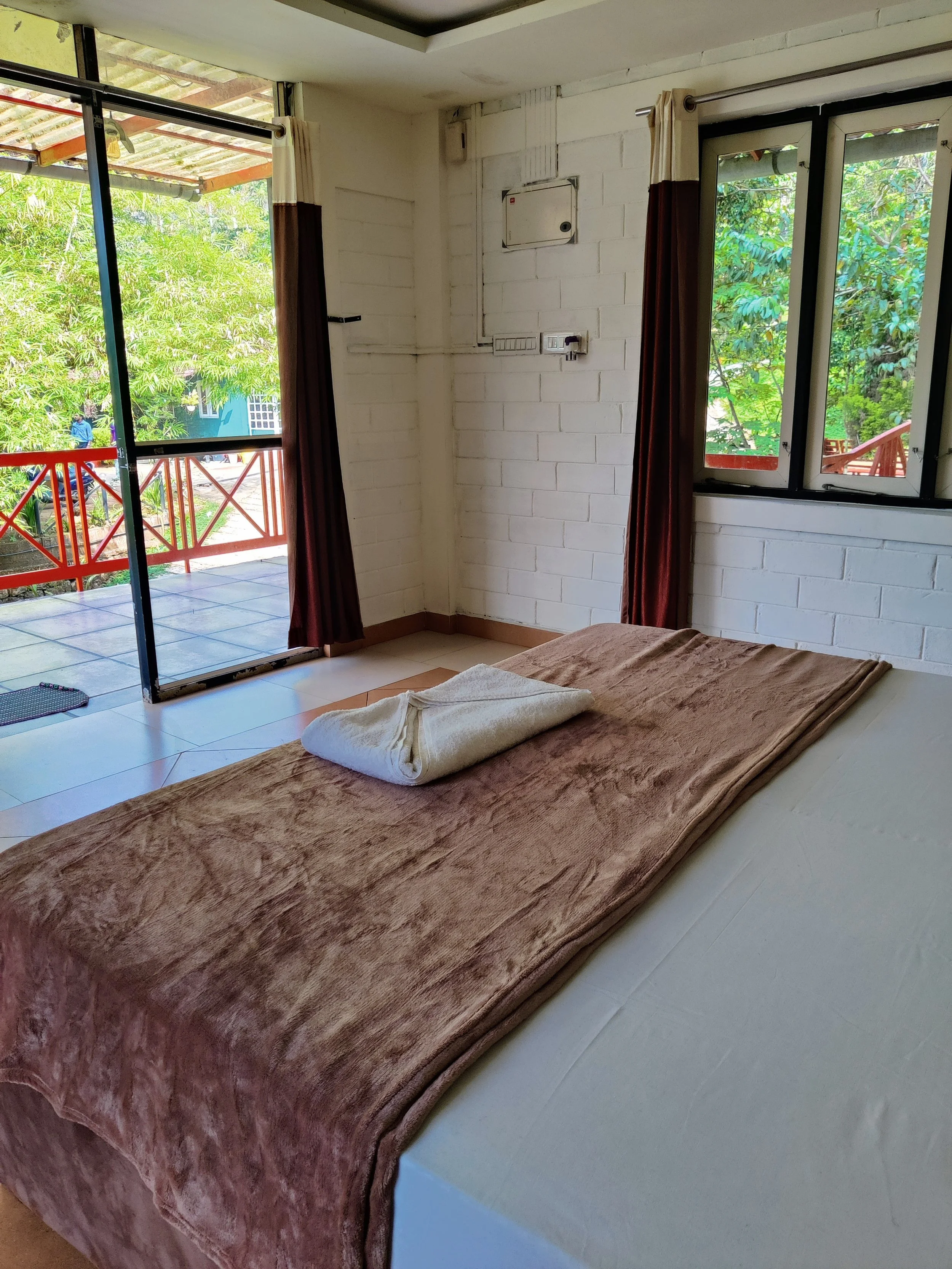 Hotel room with a bed covered in a brown blanket, towel on top, sliding glass door leading to a balcony with green trees outside, and window with white curtains.