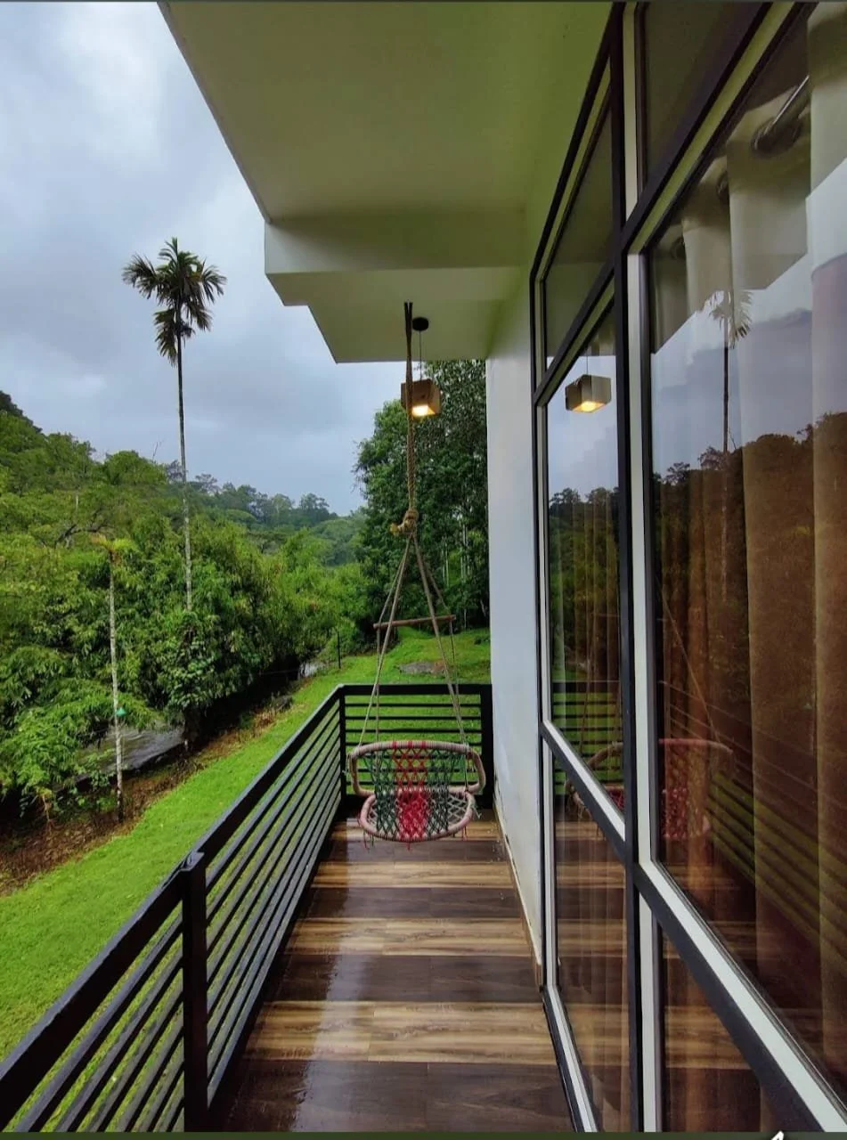 Balcony with wood flooring, black railing, hanging chair with cushion, large glass sliding doors, and lush green landscape with trees and a tall palm tree in the background.