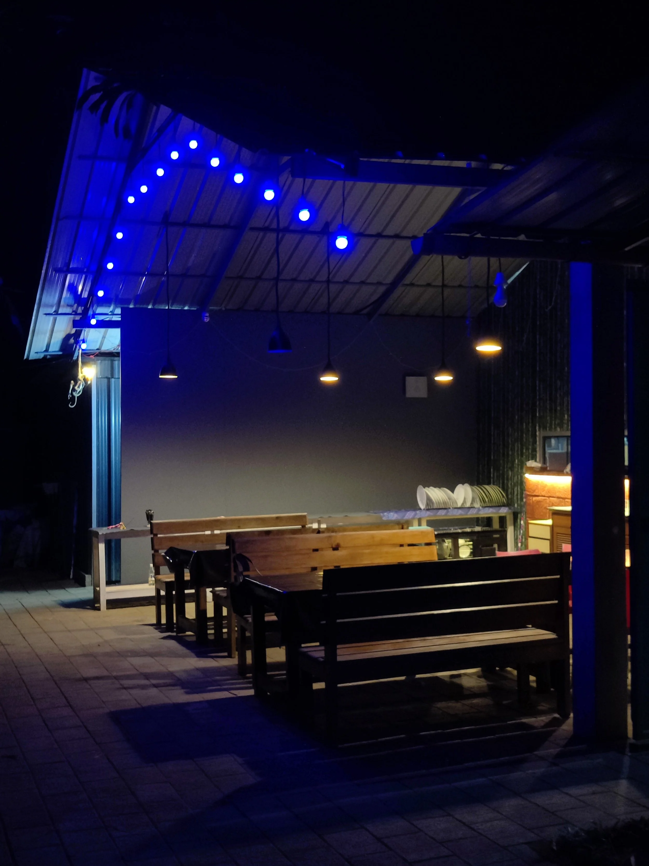 Empty outdoor seating area with wooden benches, blue string lights, and a counter with dishes, illuminated at night.