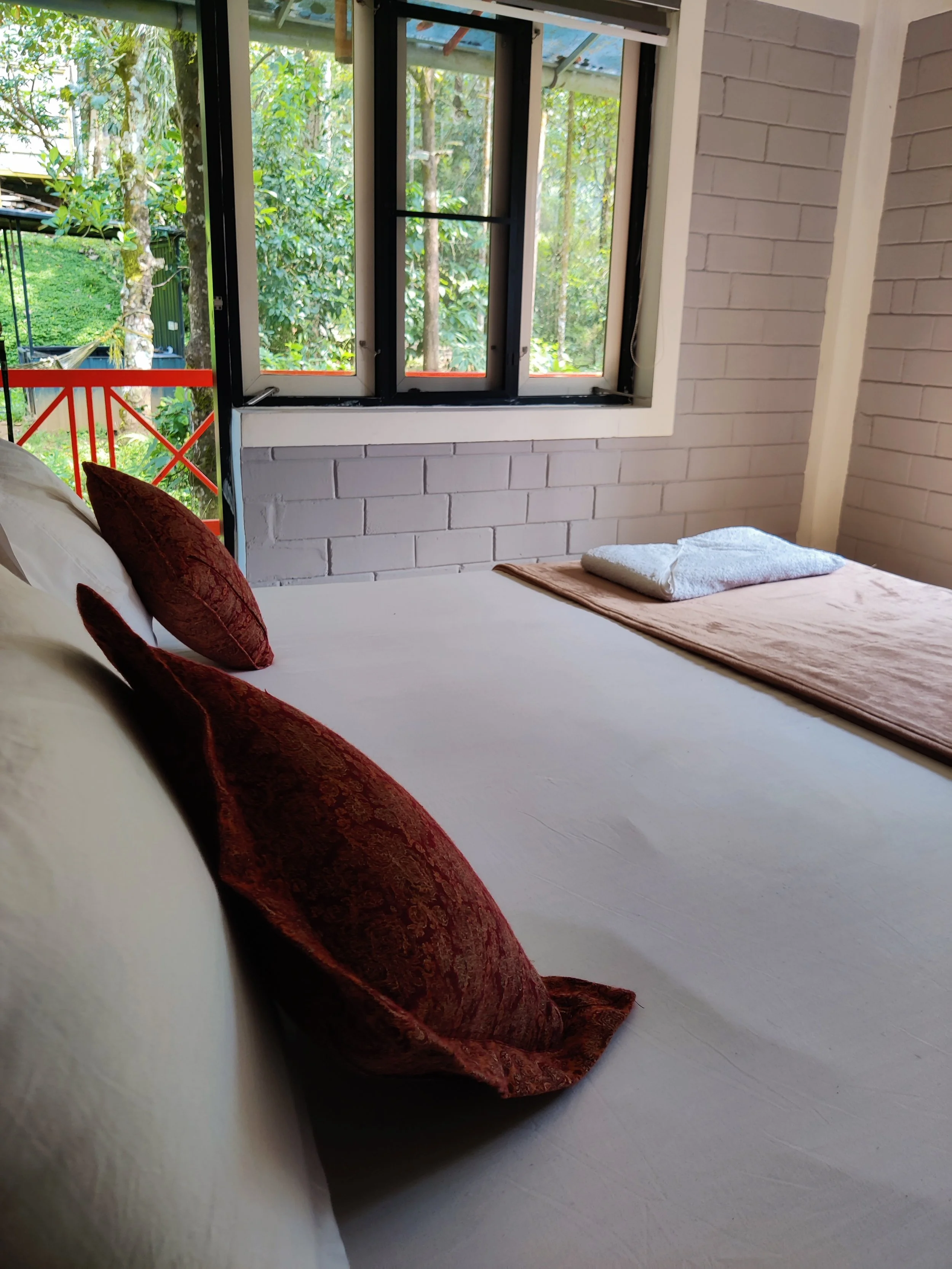 A bedroom with white brick walls and an open window showing greenery outside. There are pillows on the bed, with some towels folded at the foot.