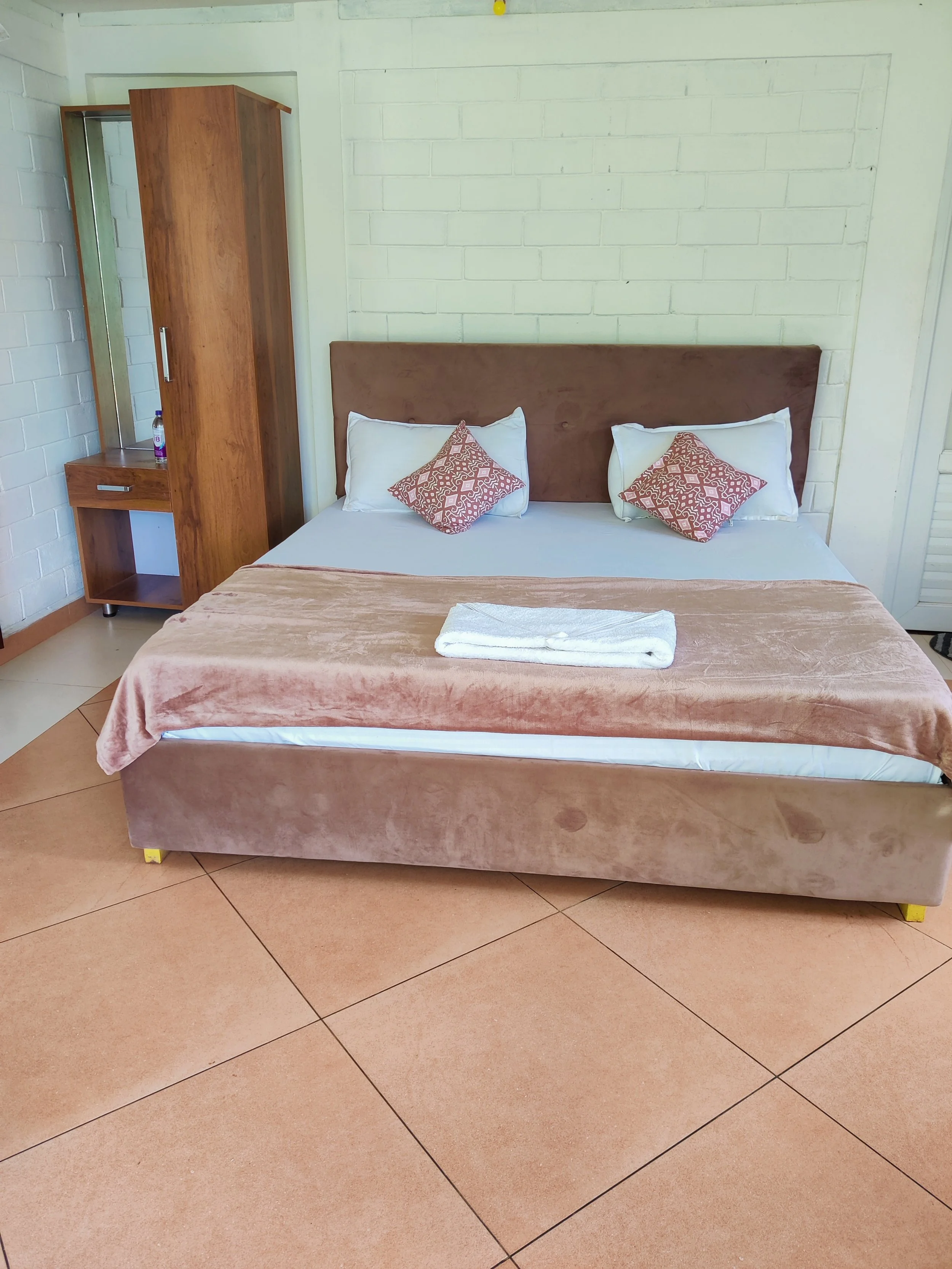 A neatly made hotel bed with a brown upholstered headboard, two white pillows with patterned red decorative pillows, a folded towel on the bed, and a wooden wardrobe with a full-length mirror in the background.
