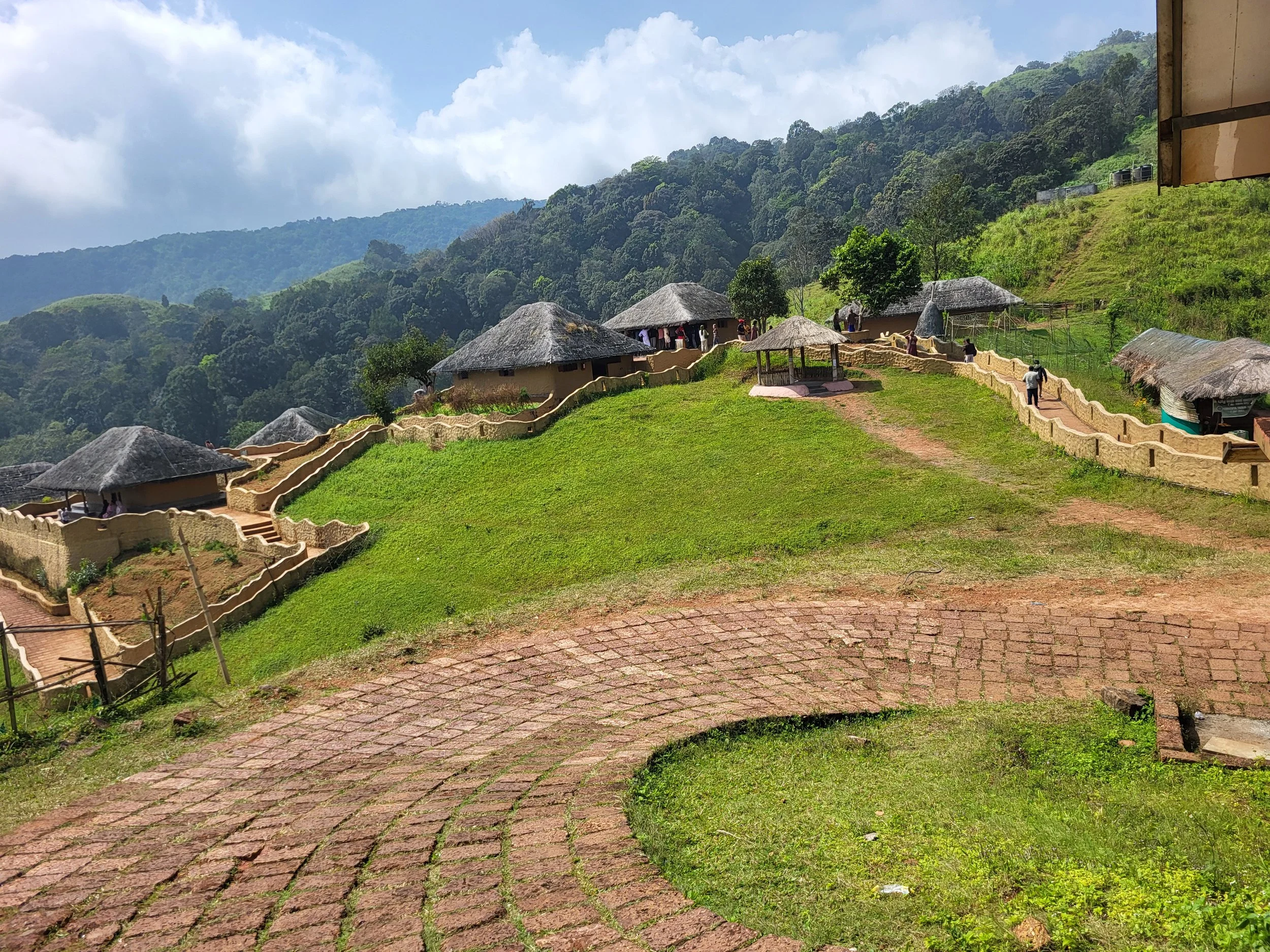 Houses with thatched roofs on a green hillside, with a winding brick path and a backdrop of forested mountains and a partly cloudy sky.