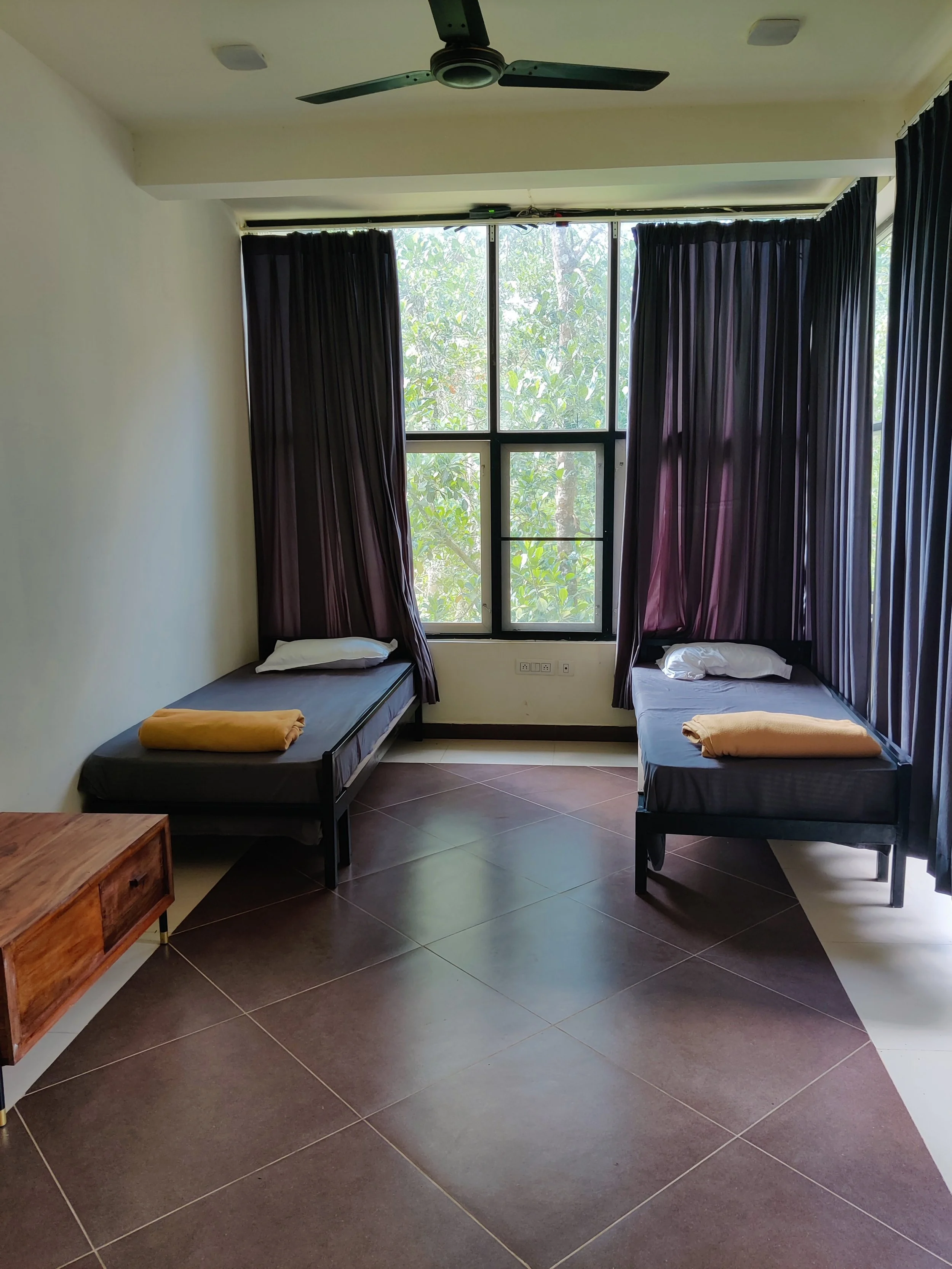 A minimalist bedroom featuring two single beds with dark linens, yellow folded blankets at the foot, and pillows. The room has a large window with purple curtains, dark tile flooring, a ceiling fan, and a small wooden side table.
