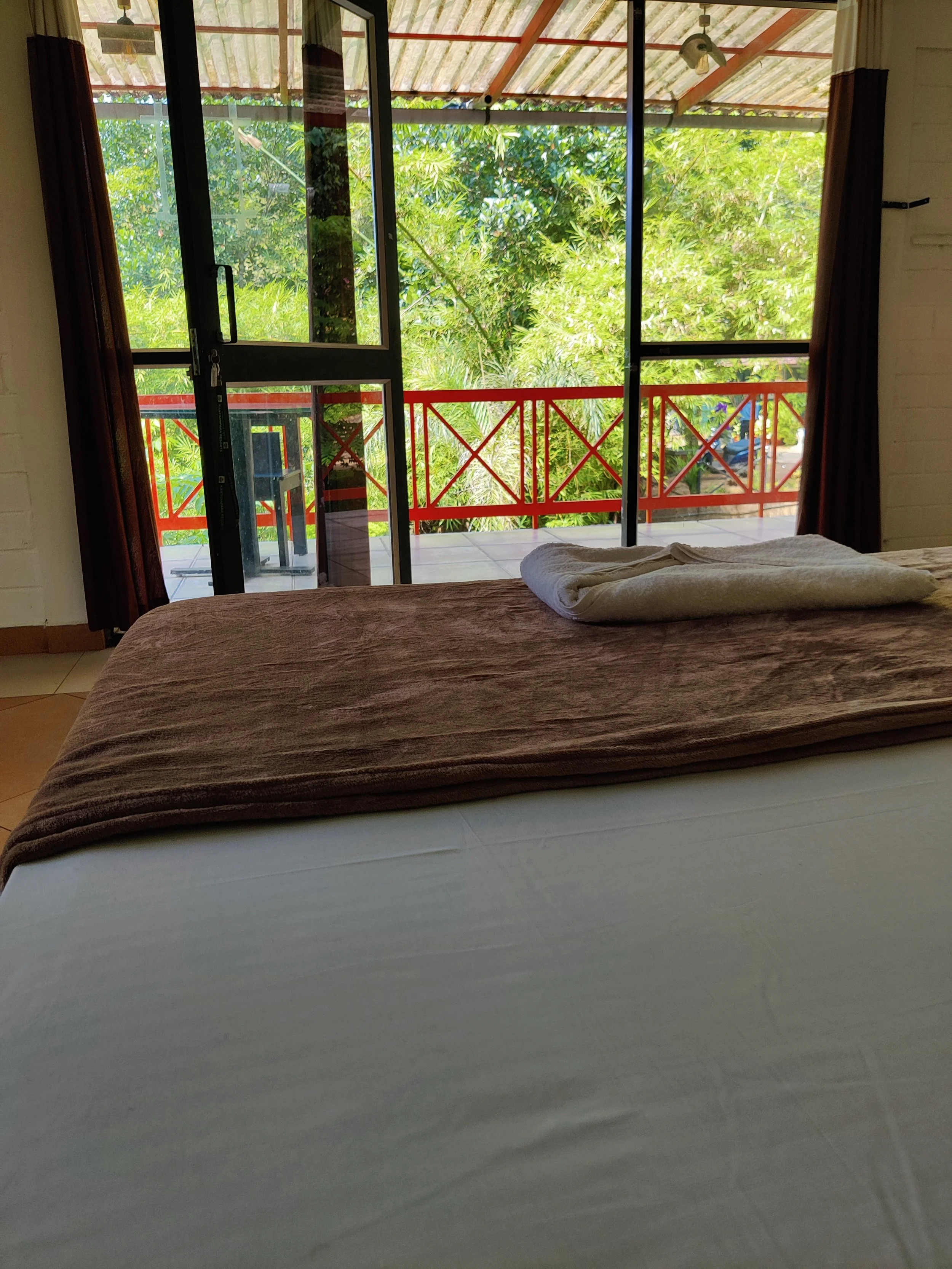 View of a bedroom with a bed, brown blanket, and folded towels, facing a glass door leading to a balcony with red railing, overlooking lush green trees.
