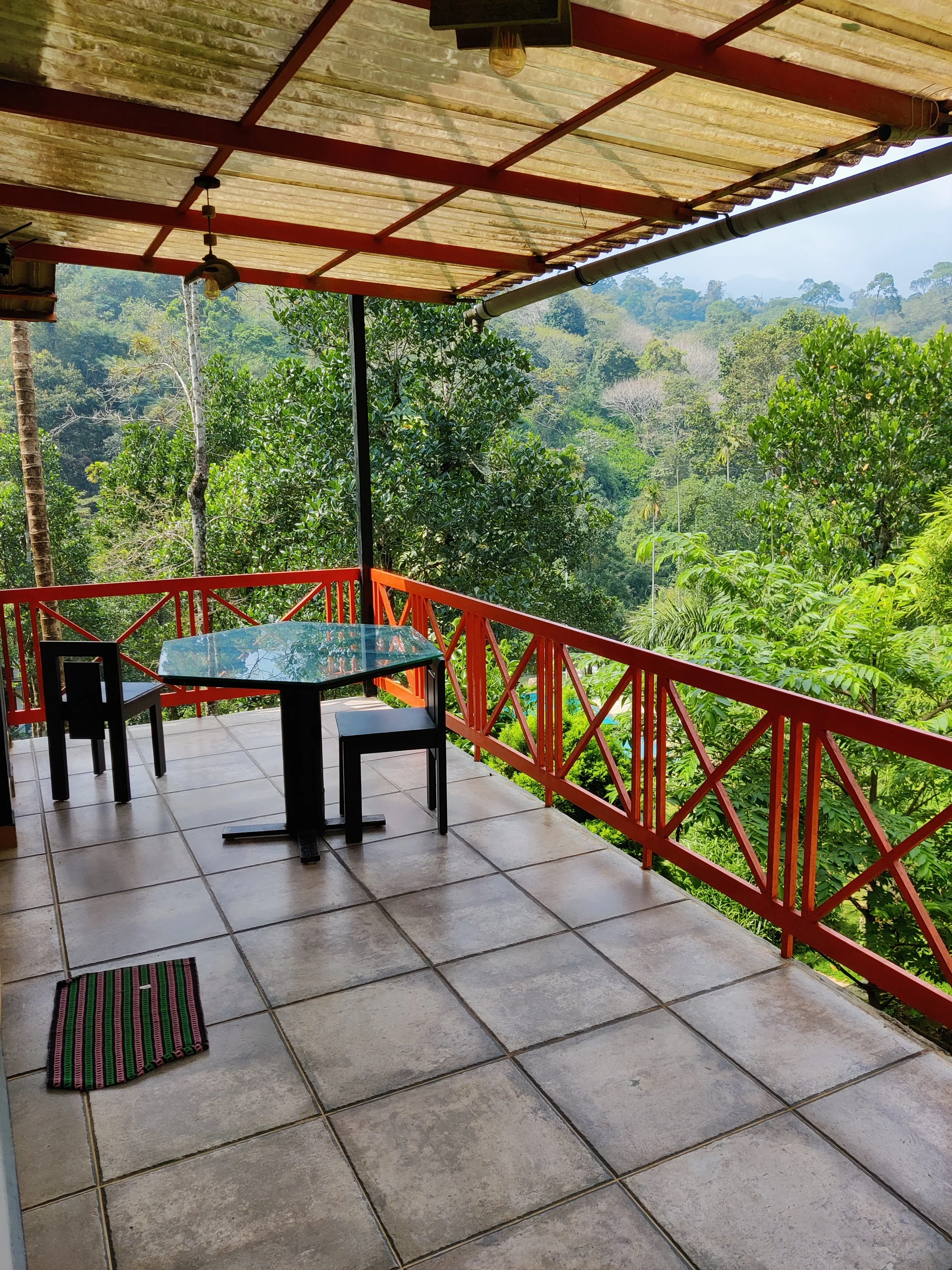 Balcony overlooking lush green forest with red railing, tiled floor, glass table, two black chairs, and a thatched roof.