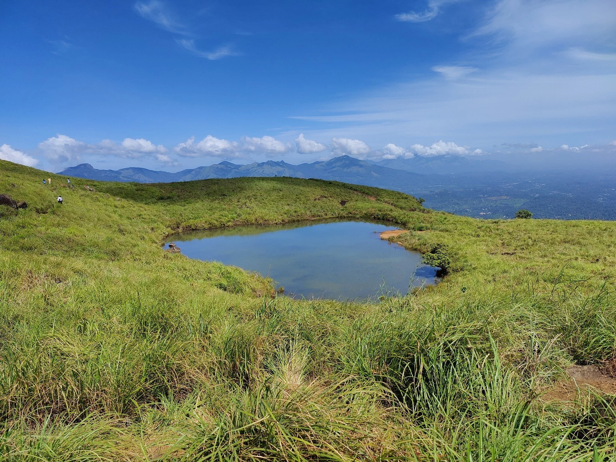 A small pond surrounded by green grass on a hillside, with mountains in the background and a blue sky with clouds overhead.