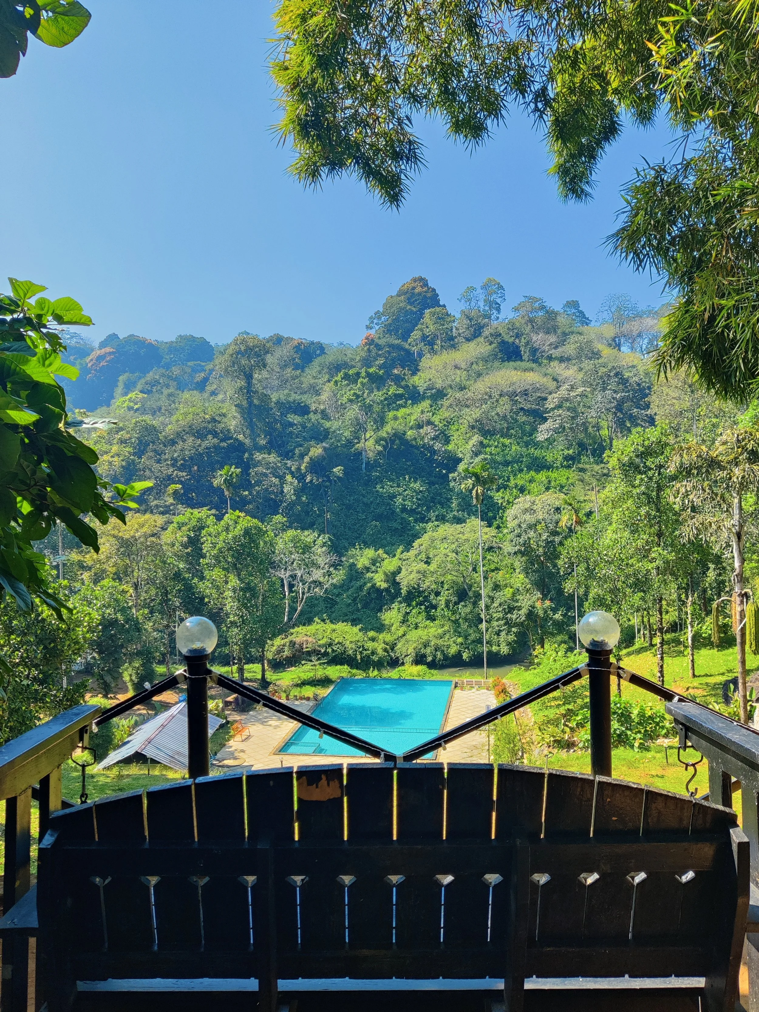 A wooden staircase with black railing and decorative posts leading down to a lush green landscape with a swimming pool surrounded by trees and mountains in the background, under a clear blue sky.