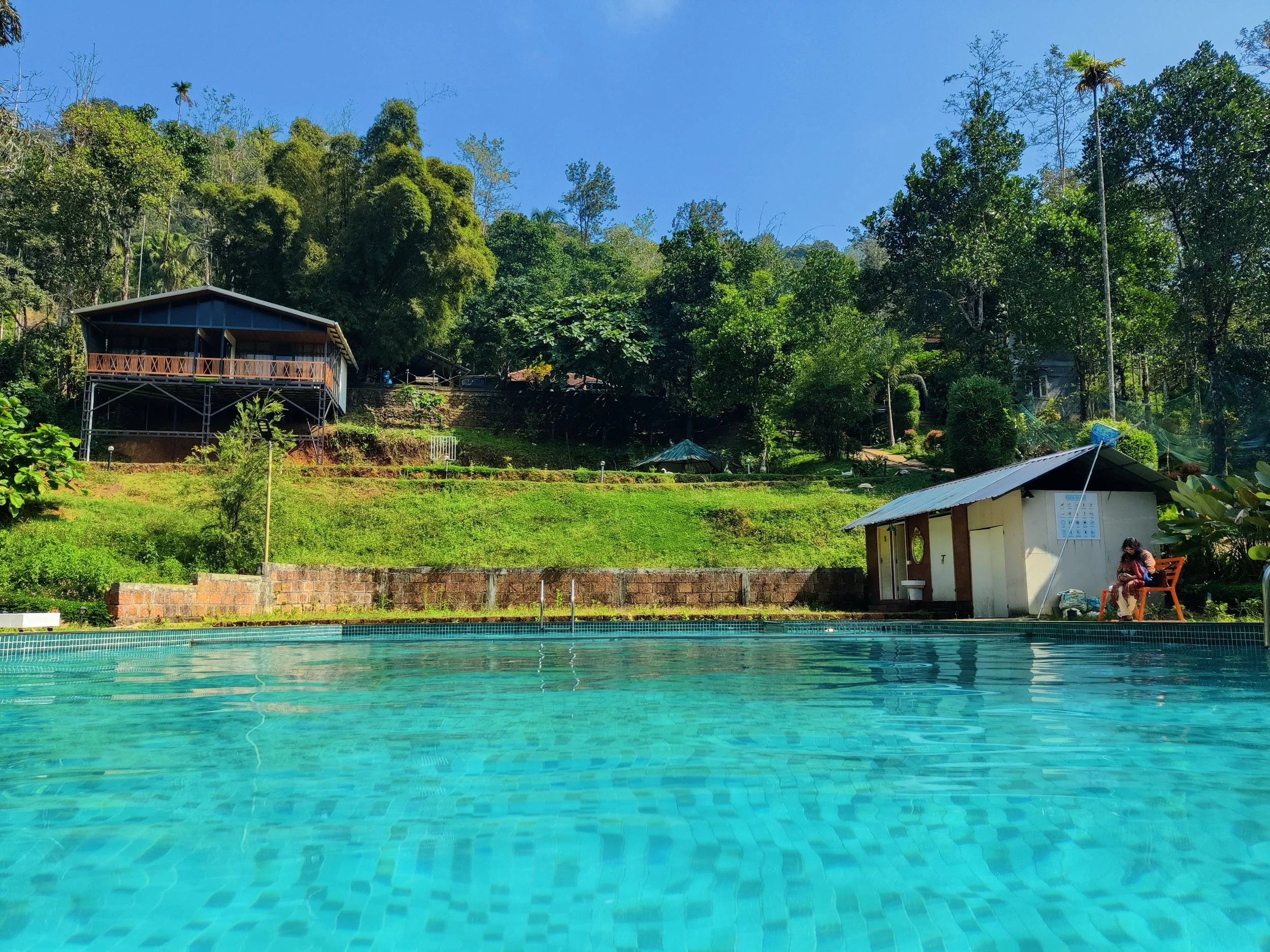 A swimming pool with clear blue water in front of a lush green hillside with trees and shrubs. There are two buildings on the hillside, one larger with a balcony and one smaller with a metal roof. A person is sitting on a bench beside the smaller bui