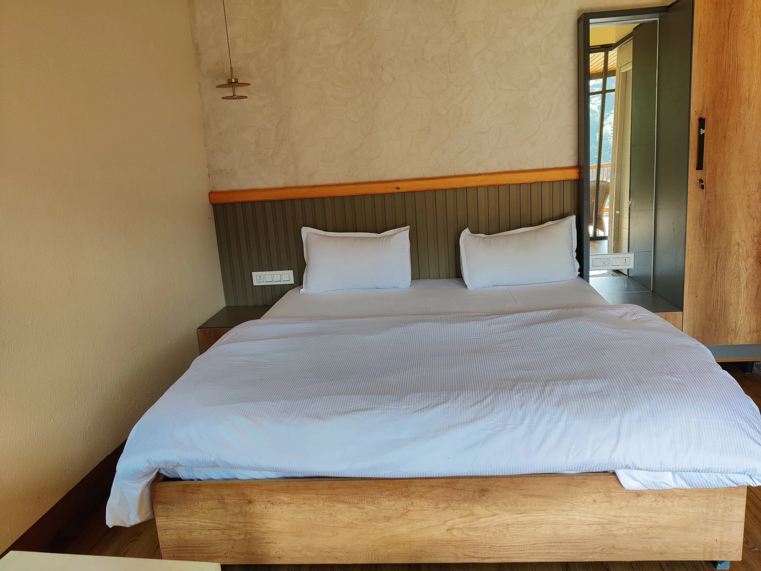 A neatly made bed with white sheets and two large white pillows. The headboard is a combination of wood and tile, with a mirror on the right side. A window with a balcony and outdoor scenery can be seen in the reflection.