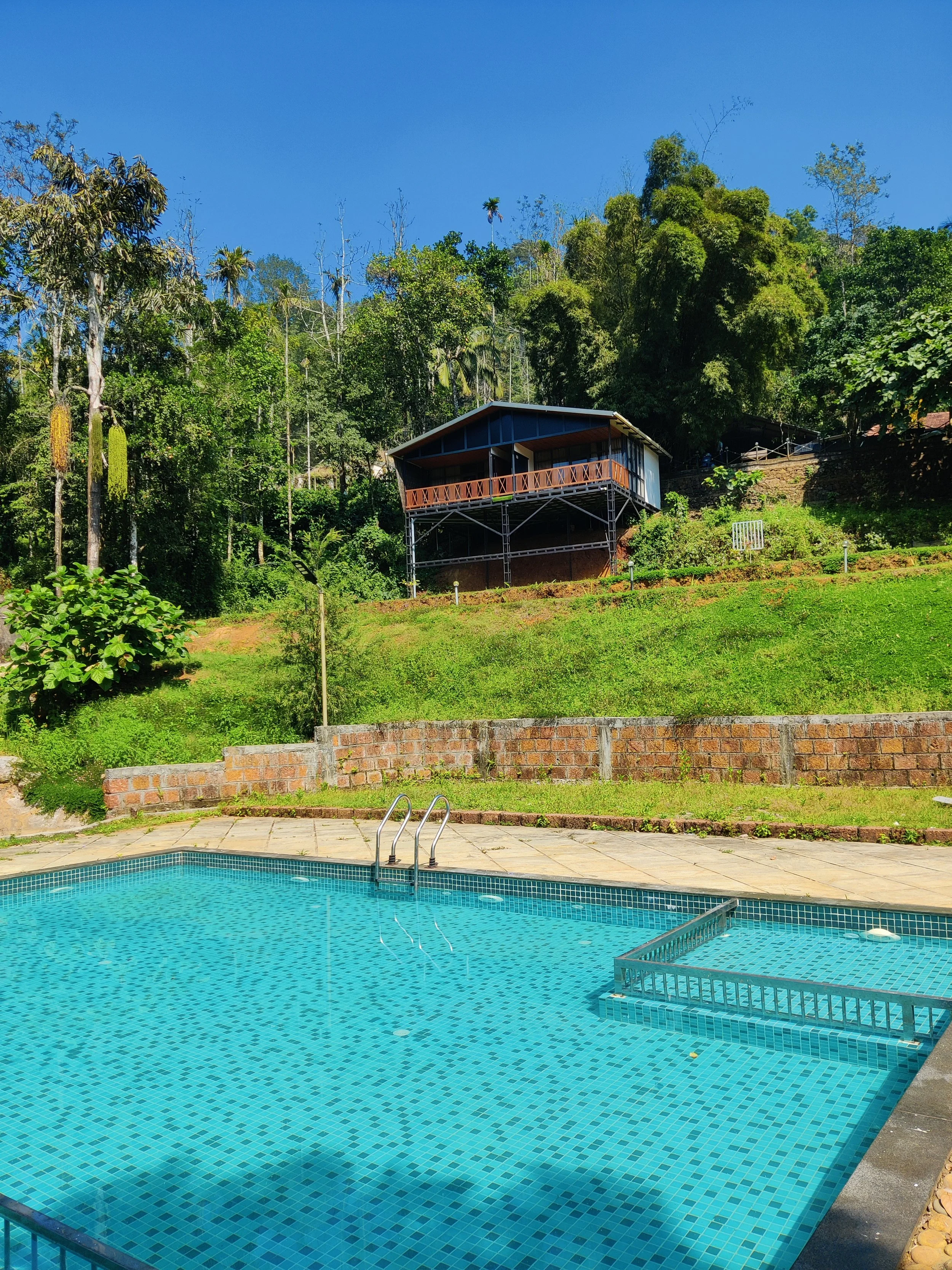 Outdoor swimming pool with metal handrail, surrounded by a paved deck, with a green hillside and a house on stilts in the background under a clear blue sky.