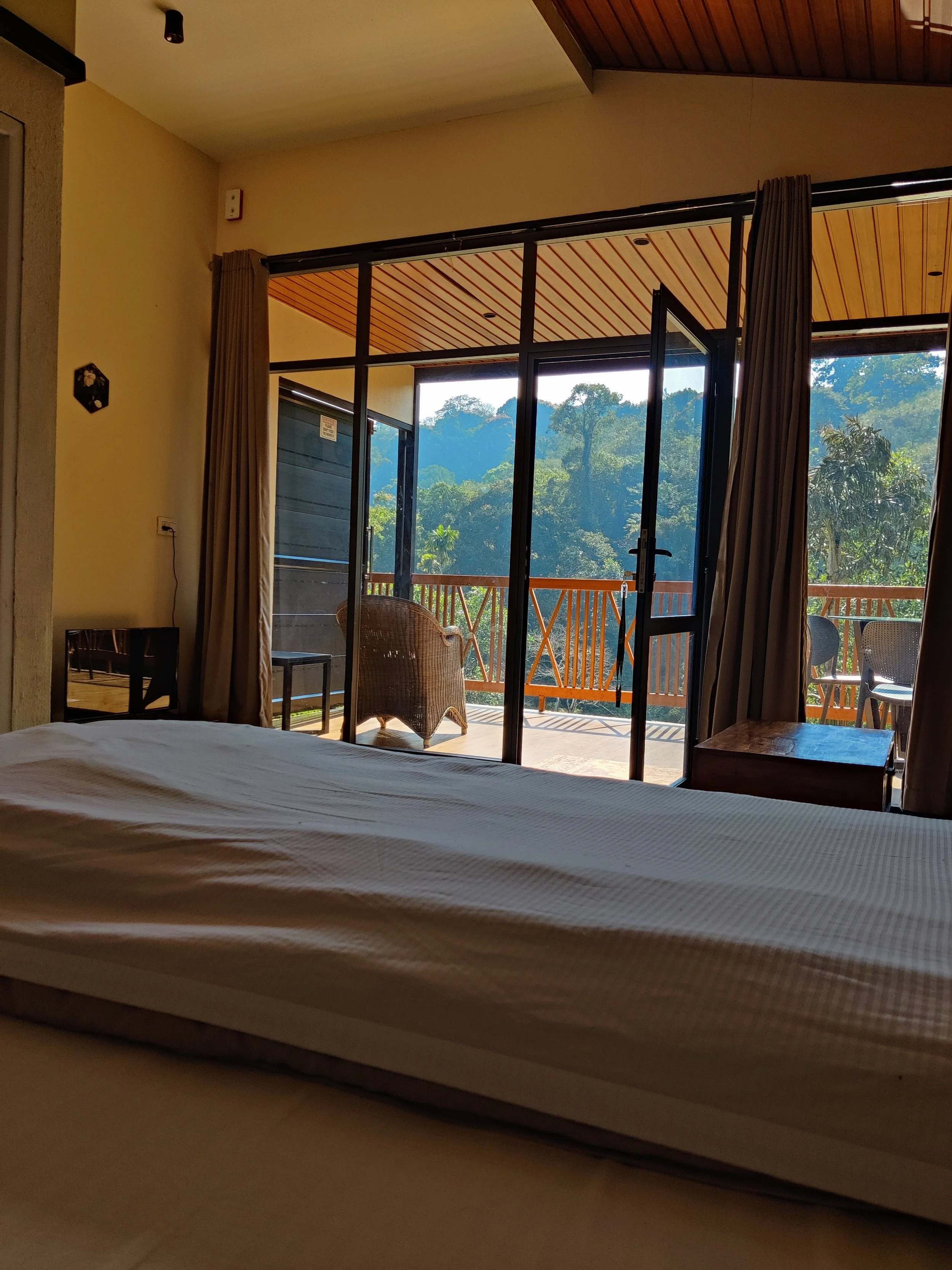 View from inside a bedroom looking out through sliding glass doors to a balcony with outdoor chairs and a scenic rainforest landscape in the background.