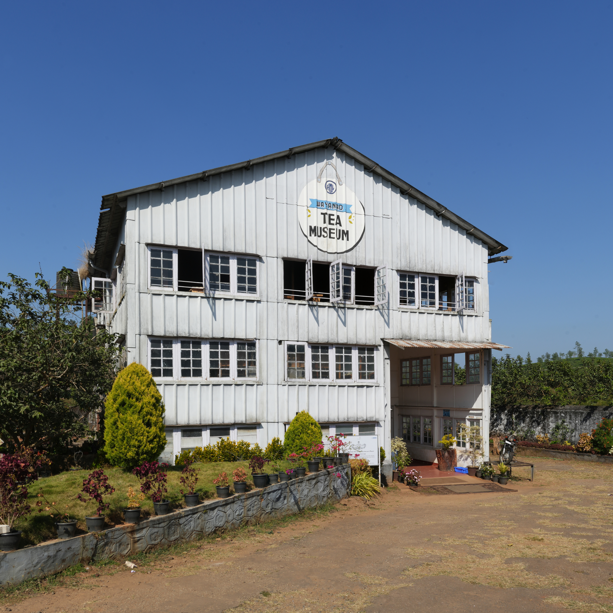 White, two-story building labeled as 'Way And Tea Museum' with open windows, surrounded by plants and flowers, under a clear blue sky.