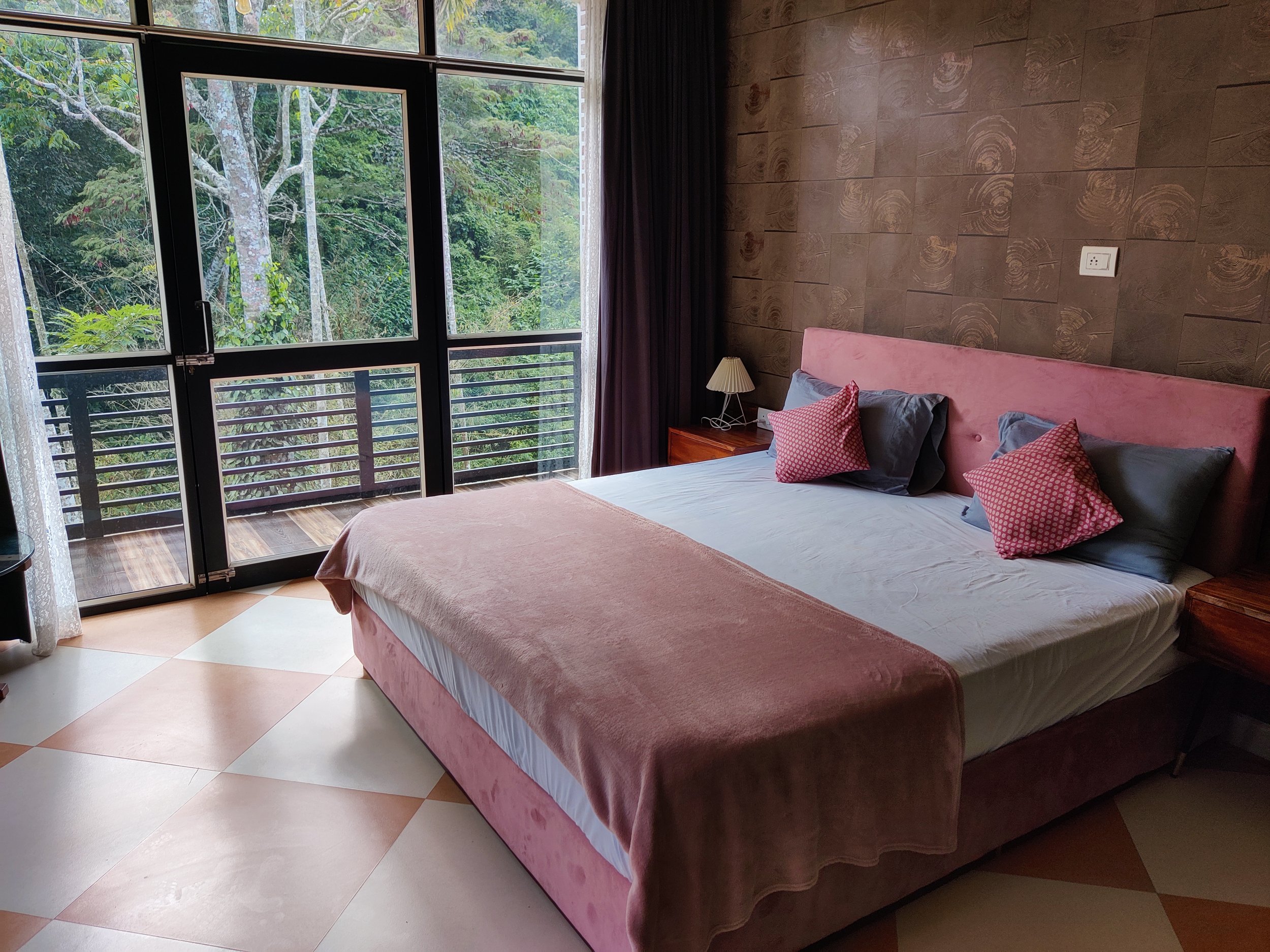 A bedroom with a large bed covered with a pink blanket, puffy pink and gray pillows, and a pink upholstered headboard. There is a small bedside table with a white lamp. The room has a glass door and large windows showing a lush, green forest outside.